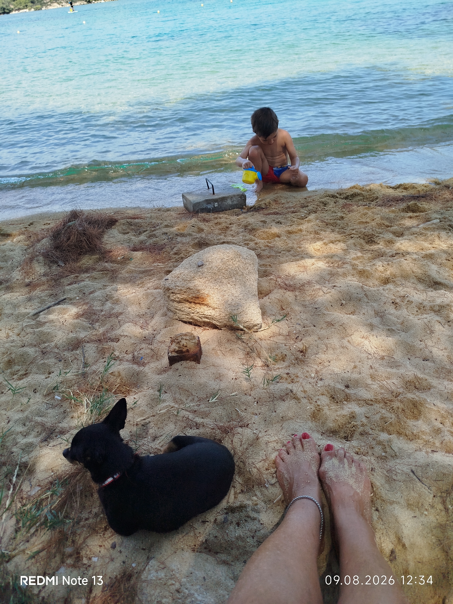 A young boy, wearing red and blue swim trunks, kneels at the edge of the turquoise sea, playing with a yellow toy bucket and a blue shovel. The boy is positioned on a sandy beach, with shallow waves lapping at the shore. To the left of the frame, a small black dog with a red and white collar lies on the sand. In the foreground, a pair of bare feet with red nail polish are visible, adorned with a silver anklet. The sand is speckled with pine needles and scattered with small stones and a large boulder. The scene appears to be a serene beach setting, likely within Sithonia Municipal Unit, Greece, given the clear blue water and Mediterranean vegetation. The image has a timestamp of "09.08.2026 12:34" and the text "REDMI Note 13" is also present.