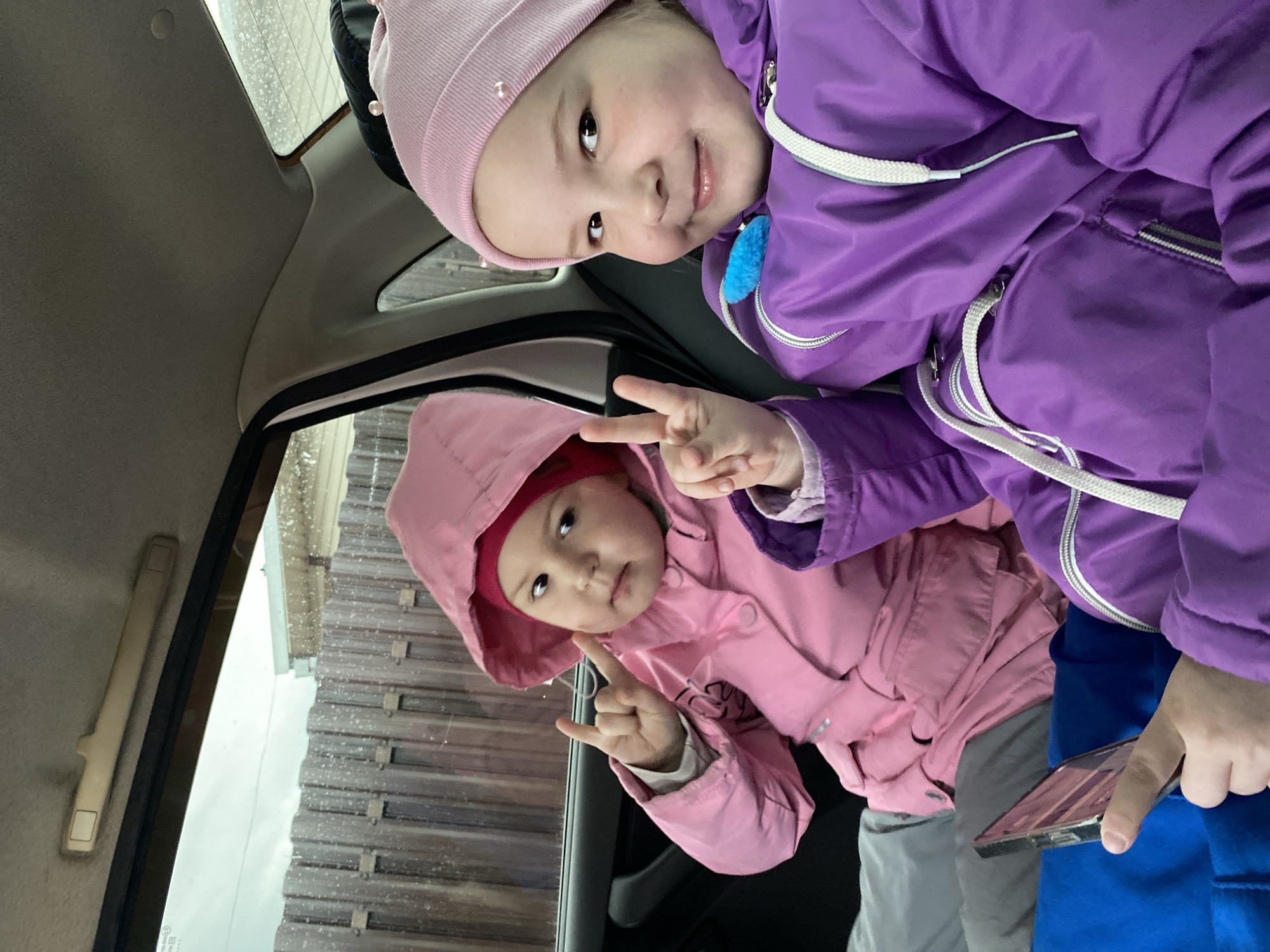 Two young children are inside a car, possibly in Chaikovsky, Russia. One child wears a pink hooded jacket and a pink hat, making a hand gesture near the car window. The other child, wearing a purple jacket and a light pink hat, smiles at the camera. The exterior of the car window shows a corrugated metal structure, possibly a building or fence, and the scene may be a car wash. The car interior is visible with neutral-colored ceiling upholstery and a handle near the window. A partially visible mobile phone is held by someone in blue trousers.