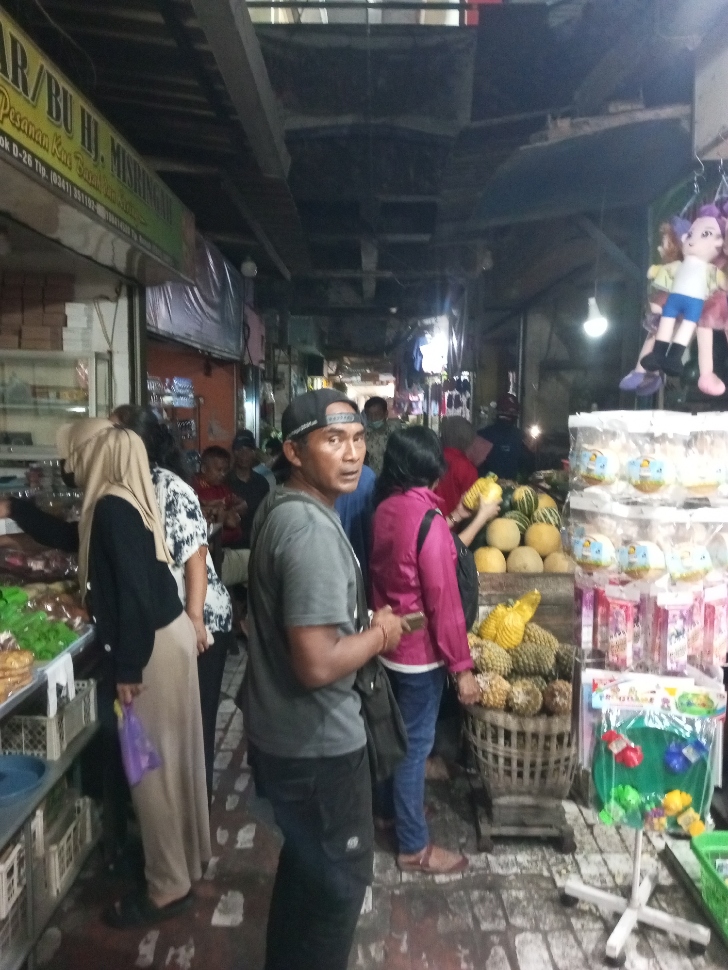 The image depicts a street market scene, likely in Malang, Indonesia, given the context of the original prompt. A man in a grey t-shirt and black cap stands in the foreground, looking to his right. Behind him, a woman in a bright pink jacket is seen reaching for fruit. To the left, a woman wearing a beige hijab and black jacket is interacting with a stall. Various fruits, including melons and what appear to be durians, are displayed in baskets. Shelves laden with packaged goods and green produce are also visible. Overhead, the market is covered by a dark, corrugated ceiling with exposed beams. Hanging from above are dolls and light bulbs, illuminating the somewhat dimly lit space. The ground is paved with cobblestones.