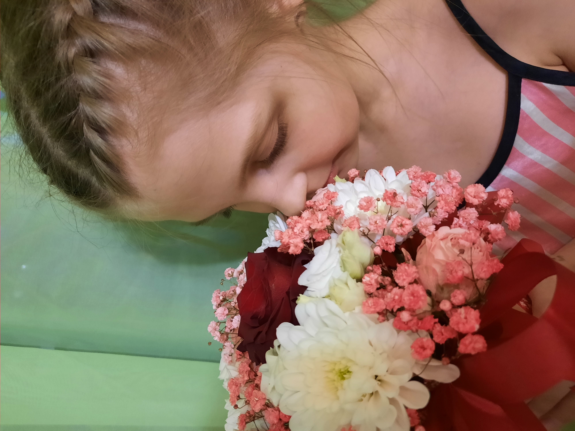 A girl with a braided hairstyle is smelling a bouquet of flowers that includes white chrysanthemums, a red rose, light pink roses, and light pink baby's breath, tied with a red ribbon. She is wearing a pink and white striped top with black straps. The background is a light green solid color. Based on available data, there is no location information available to suggest this image was taken in Izhevsk, Russia.
