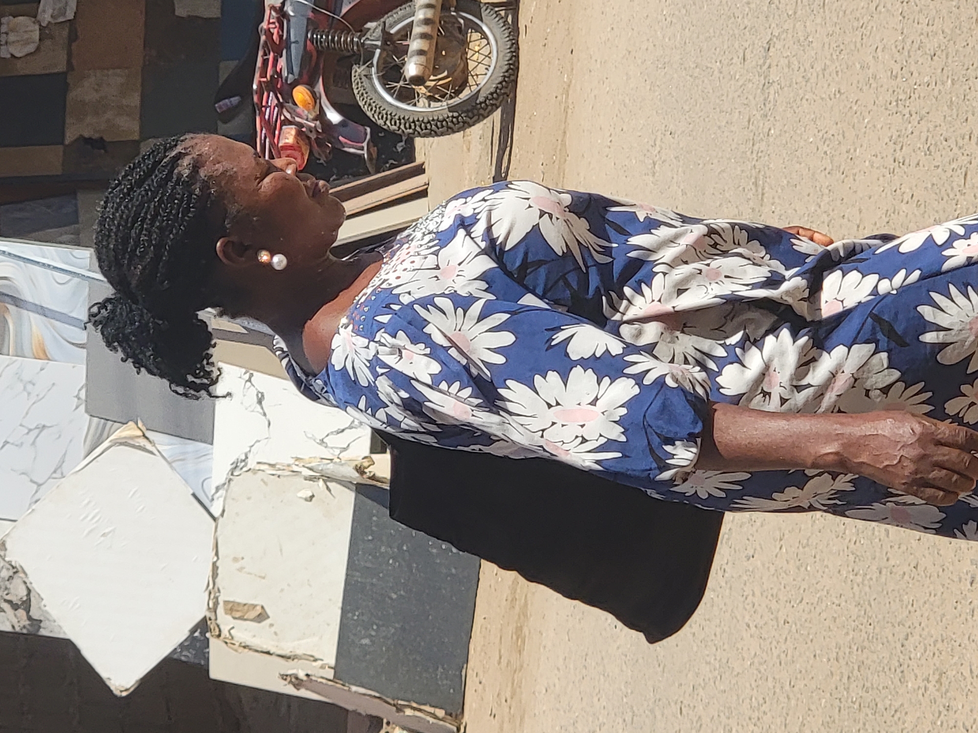 A middle-aged Black woman stands outdoors in what appears to be a semi-urban setting. She has dark, braided hair pulled back in a low ponytail and is wearing pearl earrings. She is wearing a knee-length dress with a dark blue background and white floral print. Her skin is visibly dry and veined. The ground is a pale tan color. Behind her on the left are stacked white and gray tiles and a motorcycle. The scene is in Maiburiji, Nigeria.