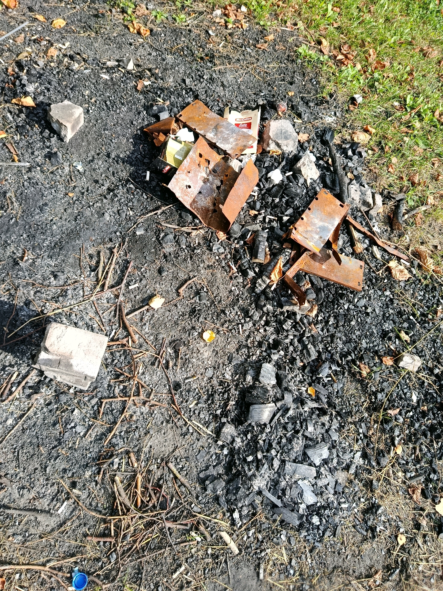 The image depicts a pile of ashes and burnt debris on the ground. Amongst the ashes are pieces of rusted metal, charcoal, a cigarette pack, and some scattered leaves. Two light-colored rectangular objects, possibly bricks or concrete pieces, are visible to the left of the pile. A blue bottle cap is on the ground in the lower left corner. The ground is a mixture of dark soil and ash, with some dry grass and twigs. The area appears to be outdoors.