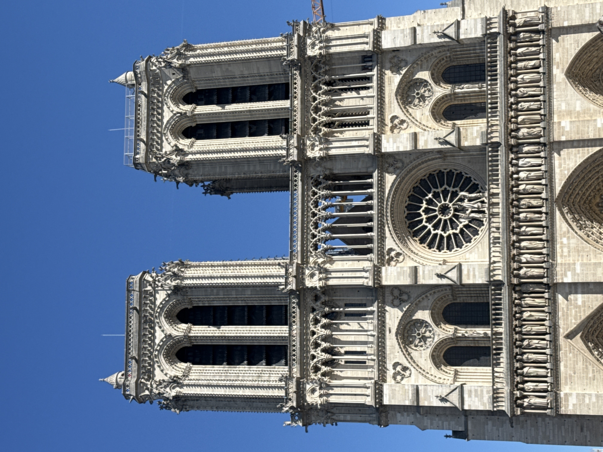 The image displays a detailed view of the upper facade of Notre-Dame Cathedral in Paris, France, set against a bright, clear blue sky. The perspective is looking upwards, emphasizing the grandeur and intricate Gothic architecture.
The central feature is the cathedral's iconic twin towers, both heavily decorated with archways, pinnacles, and carved details. Between the towers, a large, ornate rose window with dark tracery is prominent, flanked by smaller decorative windows and a horizontal gallery above it. Below the rose window, the Gallery of Kings is visible, featuring a row of numerous carved statues of figures, likely royalty or saints, set within architectural niches. Further decorative elements, including arches, columns, and projecting gargoyles, adorn the entire stone structure. The stone appears weathered yet well-maintained, highlighting centuries of craftsmanship.
Visible signs of ongoing restoration or construction are present. On the left tower, white scaffolding is erected, extending upwards. In the upper right portion of the image (when oriented vertically), part of an orange construction crane can be seen peeking over the top of the structure, further indicating work in progress following the 2019 fire. A faint white contrail from an aircraft is also discernible in the vast blue sky.
The scene depicts a sunny day, likely mid-morning or afternoon, with strong natural light illuminating the cathedral's intricate details and casting subtle shadows. There is no visible text in the image. The overall impression is one of historical majesty, resilience, and ongoing preservation efforts.