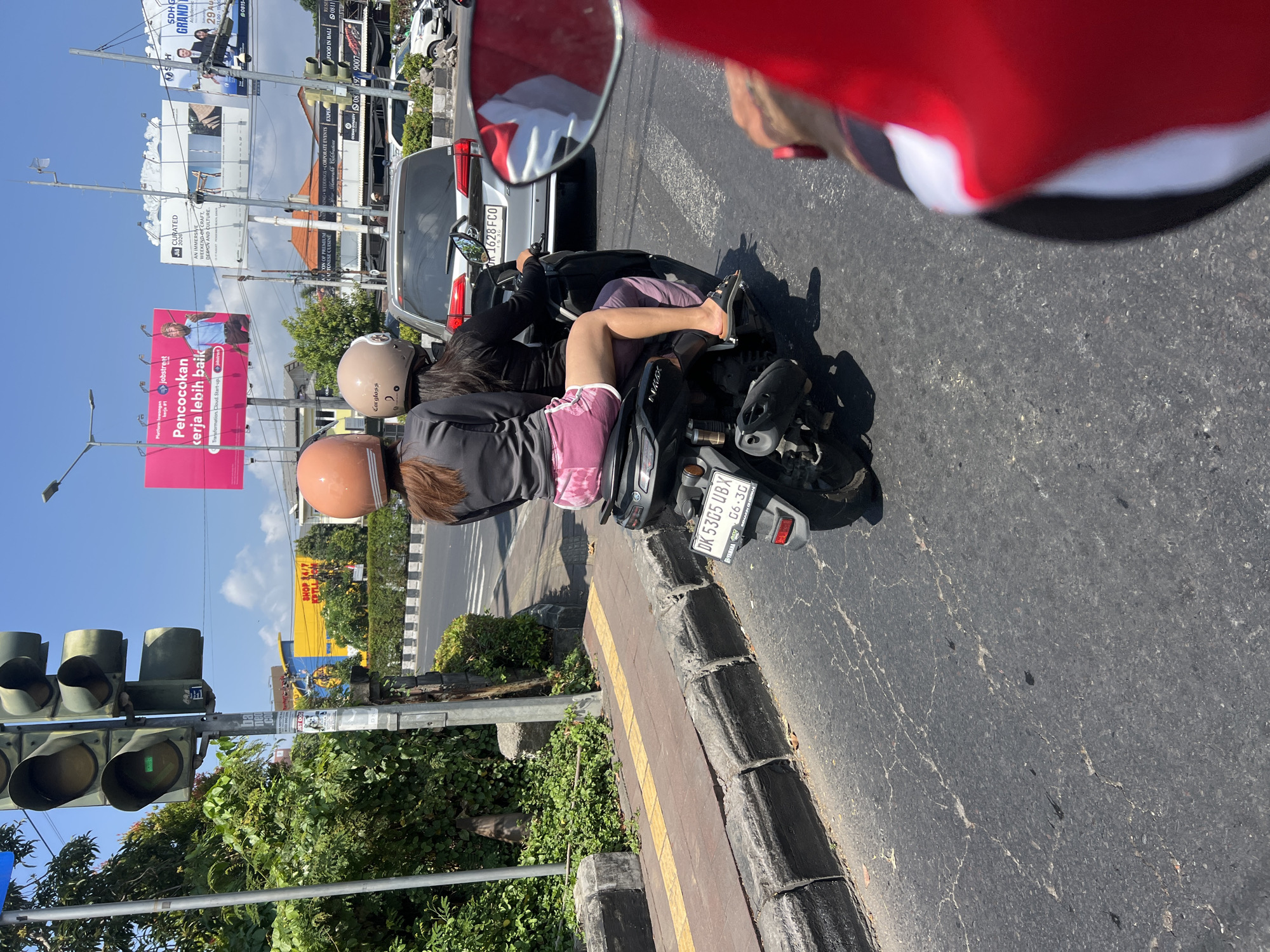In Denpasar Barat, Indonesia, two individuals are riding a scooter on a sunny day. The person in the front is wearing a light brown helmet, and the person in the back is wearing an orange helmet. The passenger is wearing pink shorts and a black long-sleeved shirt. The scooter's license plate reads "DK 5305 UBX". In the background, a silver car with the license plate "B 1628 FCO" is visible, along with billboards advertising various services. The scene takes place on a paved road with a sidewalk and curb on the left side. Traffic lights are visible in the upper left corner of the image. The overall environment suggests a busy urban street.