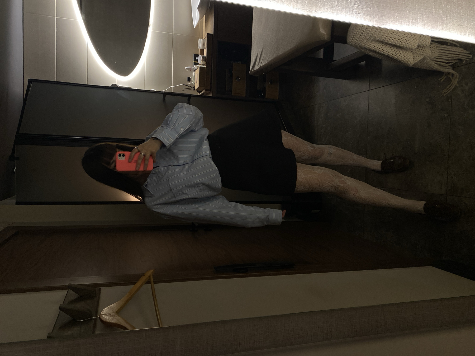 A person wearing a blue striped shirt, black skirt, and white patterned tights is standing on a gray tile floor. They are holding a pink phone up to their face, possibly taking a picture. The room includes a dark desk or cabinet, a bed or seating area with a textured throw, a light fixture, and a wooden hanger in the foreground. The exact location is not discernable from the image.