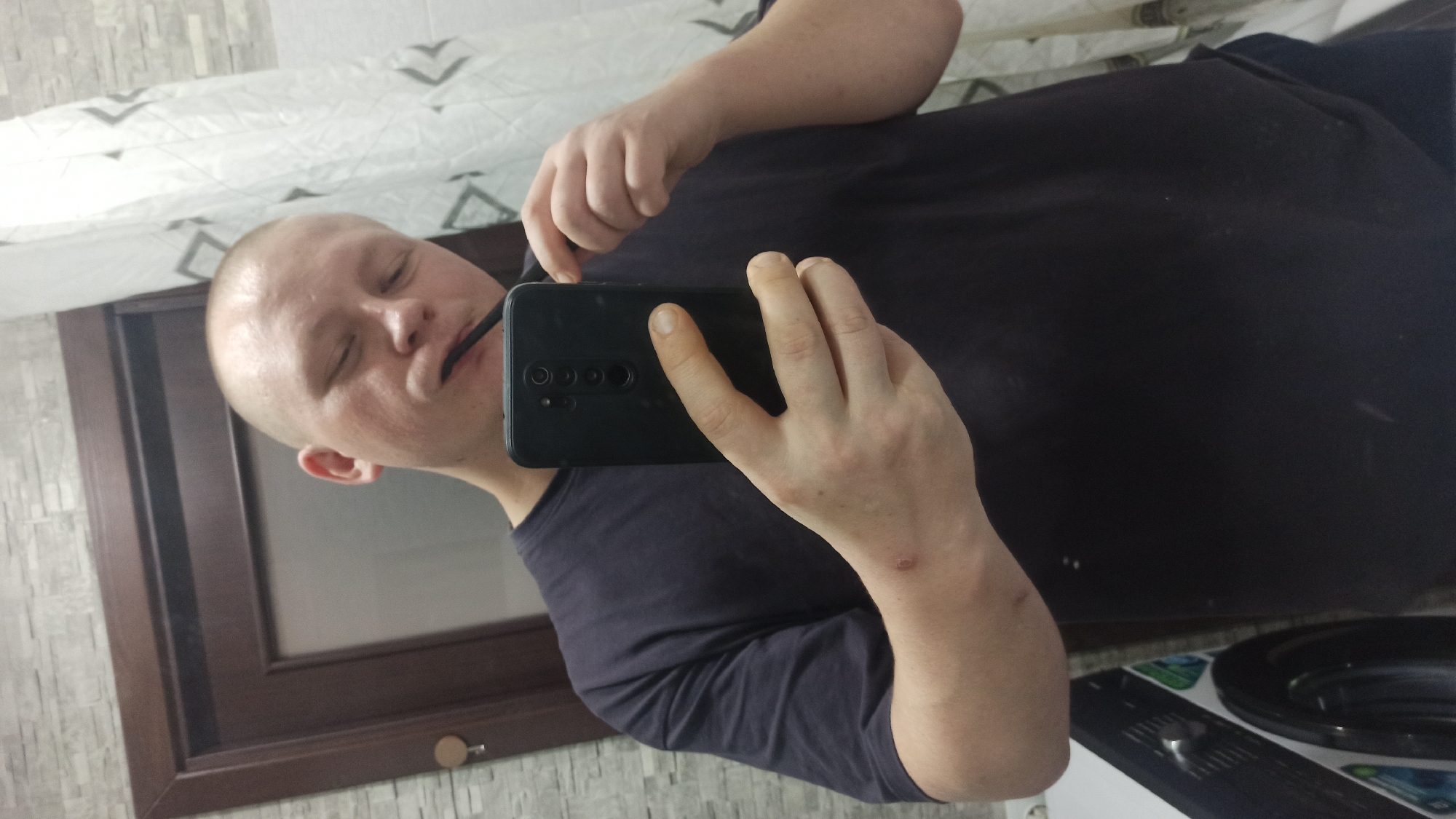 In an indoor setting, possibly a bathroom, a bald, fair-skinned individual is holding a smartphone to take a selfie while brushing their teeth. The person is wearing a dark t-shirt and their left arm is visible, with a visible wound or mark on the forearm. The background shows a tiled wall, a wooden door, and a shower curtain with a geometric pattern. To the left, a portion of a washing machine is visible. The lighting appears to be from an overhead source. The overall scene depicts a mundane, everyday activity captured in a mirror.