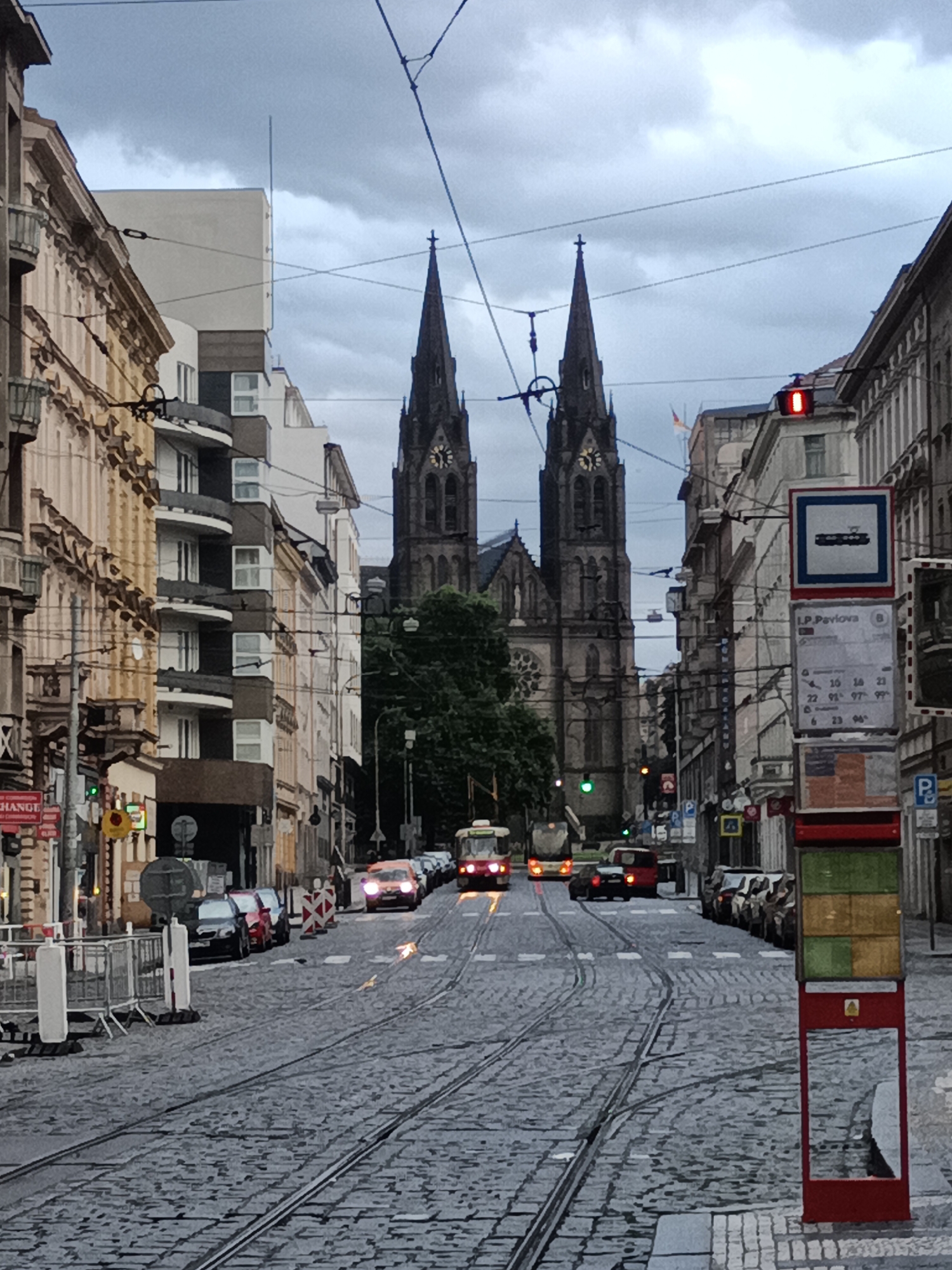 The image depicts a street scene in Prague, Czechia, with a prominent view of the Church of Saints Peter and Paul, located in Vyšehrad. The church's twin spires dominate the background. The foreground shows a cobblestone street with tram tracks. Several trams and cars are visible on the street. The weather appears overcast. A bus stop sign is visible on the right side of the image. The architecture of the surrounding buildings is typical of Prague, with ornate facades and balconies.