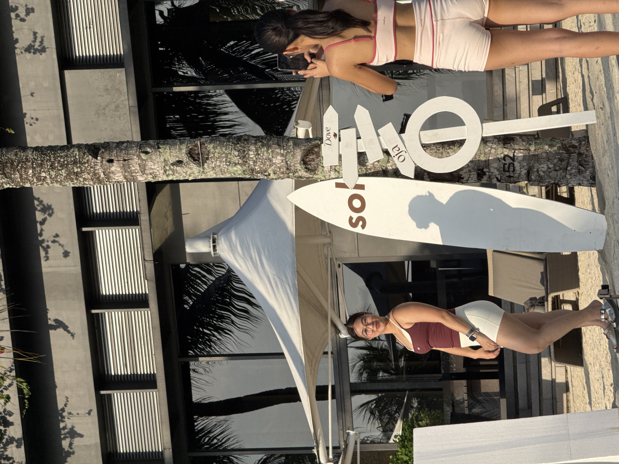 The image depicts a tropical outdoor scene, possibly at a resort or beachside establishment. In the foreground, two young women are present. The woman on the left is facing the camera, posing. She has dark hair pulled back, is wearing glasses, and is dressed in a maroon tank top and white shorts. She is standing on sand, wearing sandals.
To the right of the first woman, a palm tree stands prominently. Attached to the palm tree are several white directional signs, some with text. One sign clearly reads "Dove," and another reads "oja." A large white surfboard is leaning against the palm tree on the left side, with the word "sok" visible on it. Behind the surfboard and palm tree, there is a white tent-like structure.
Further in the background, a modern building with reflective windows and horizontal blinds is visible. Palm tree fronds are reflected in the windows. The overall setting suggests a sunny day with clear skies. The image appears to be taken in Samal, Philippines, given the presence of palm trees, the tropical climate suggested by the attire, and potentially the architectural style which is common in resort areas of the Philippines.