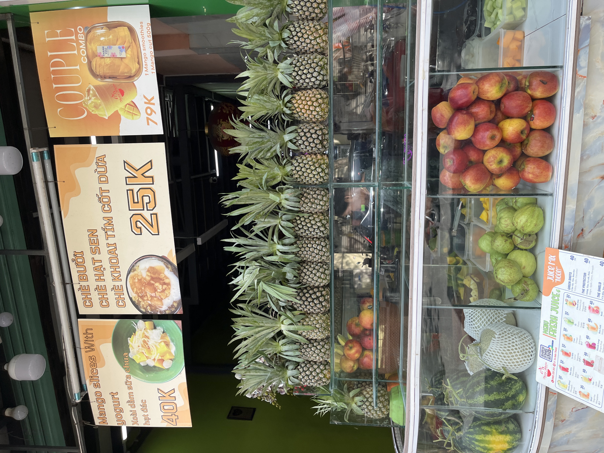 A street-side juice stall in Nha Trang, Vietnam, displays a variety of fresh fruits and pre-packaged beverages. Three illuminated signs hang above the stall. The left sign advertises "Mango slices With yogurt" for 40K, featuring an image of mango slices in yogurt. The middle sign promotes "Chè bưởi, Chè hạt sen, Chè khoai tím cốt dừa" for 25K, accompanied by a picture of a dessert. The right sign highlights a "COUPLE COMBO" for 79K, showcasing a mango smoothie and a container of sliced mango. Below the signs, a row of fresh pineapples is arranged on a shelf. Further down, glass display cases reveal an assortment of fruits, including watermelons, melons, apples, and guavas, as well as containers of cut fruits. A laminated menu, titled "FRESH JUICE", is positioned on the counter, listing various juice options and their prices. The overall environment suggests a busy urban setting, typical of a marketplace or commercial street.