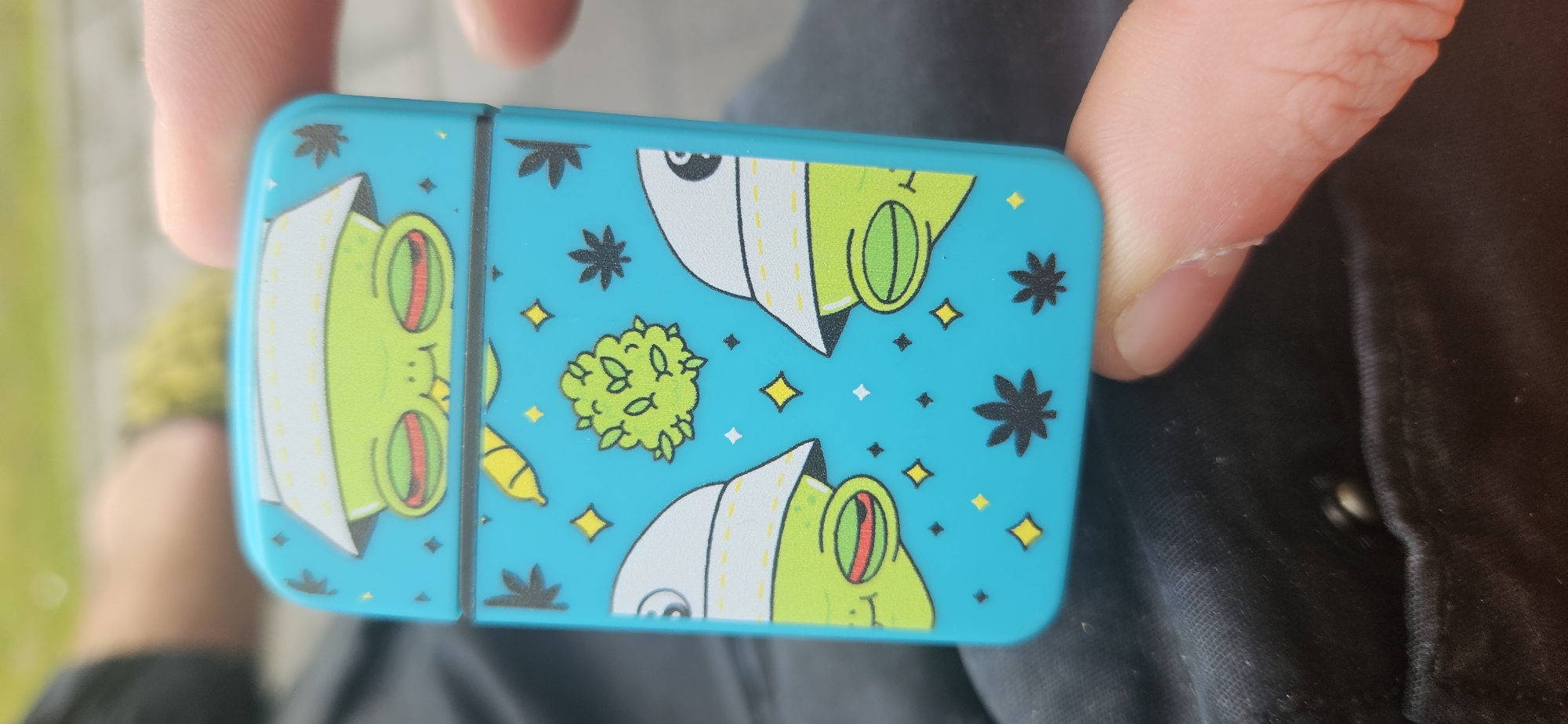 The image shows a person holding a blue metal container with a cartoon frog design. The design includes green frogs in white jackets, cannabis buds, stars, and marijuana leaves on a light blue background. The container appears to be in the hands of an individual wearing a black shirt. The location in the city of Plovdiv, Bulgaria, is not discernible from the image.