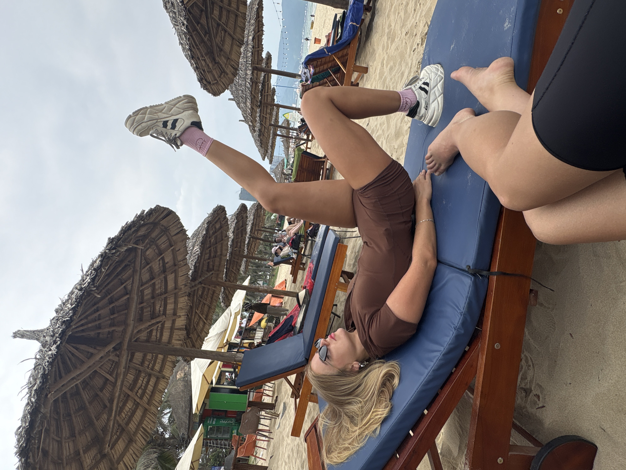 This image captures a relaxed beach scene in Da Nang, Vietnam, taken from a slightly tilted perspective. The setting features light sandy beaches, numerous large thatched umbrellas, and blue lounge chairs.
The primary subject is a person, likely a young woman, with long blonde hair, lying on her back on a blue lounge chair in the foreground. She is wearing a brown short-sleeved top and matching shorts. Her legs are raised high, with her left foot prominently visible against the sky. She wears white chunky sneakers with black stripes and light pink ankle socks that have faint white lettering, possibly "Love." Sunglasses are perched on her face, and a silver bracelet is on her wrist. She appears to be relaxing, possibly gazing upward.
To the right of the main subject, a partial view of another person's legs, clad in black shorts, is visible on an adjacent blue lounge chair. In the mid-ground and background, more people are seen lounging under similar umbrellas on the beach, with the ocean visible further out. Distant mountains or hills line the horizon under a partly cloudy sky. The overall atmosphere is one of leisure and vacation. A black zip tie can be seen securing the cushion to the wooden frame of the lounge chair in the foreground. The image depicts bright daylight conditions.