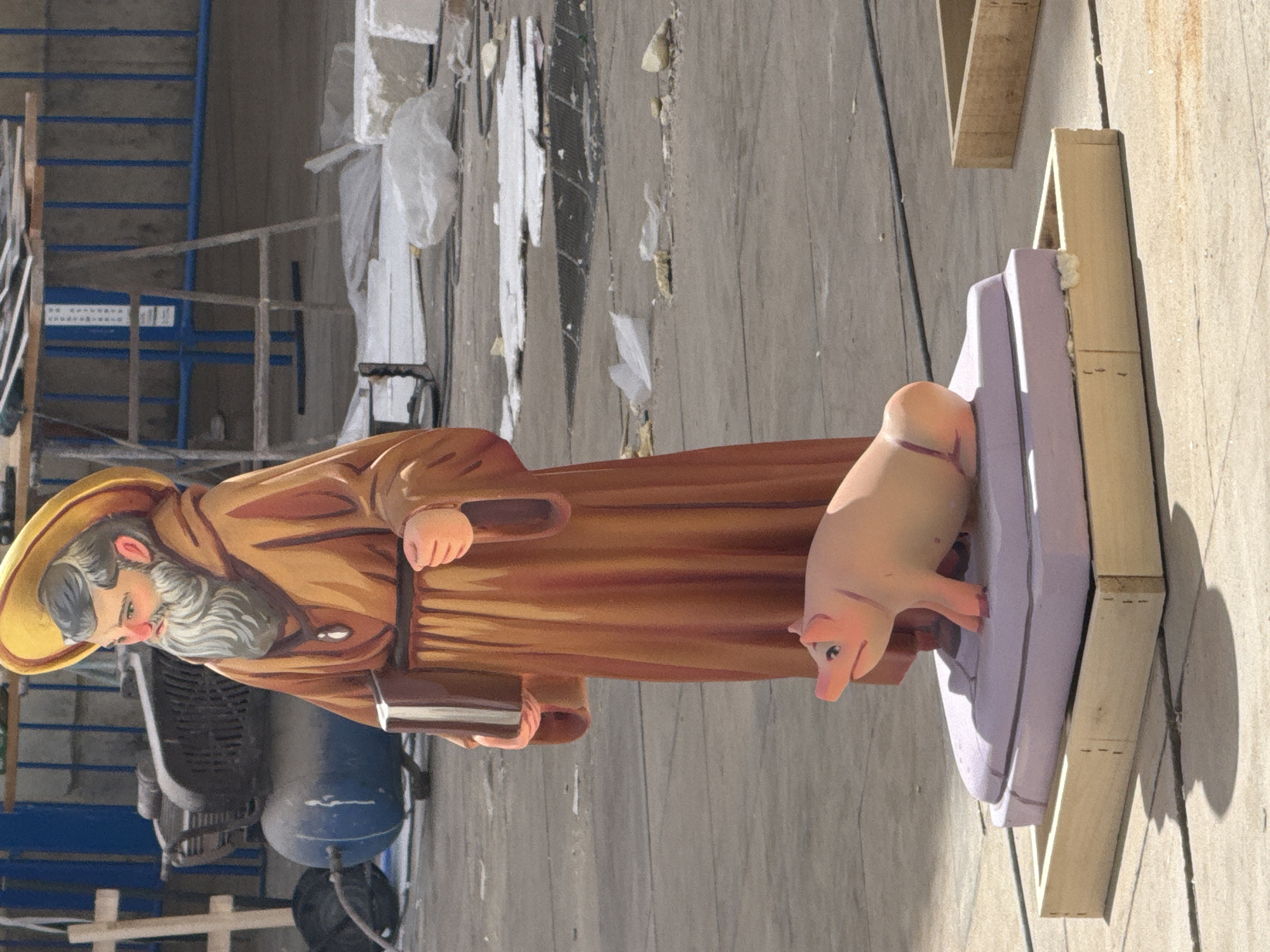 A life-sized, brightly painted statue of a bearded man in a brown robe and yellow hat stands in the foreground. He is holding a dark book in his left hand. In front of the man's statue, a smaller statue of a pink pig is perched on a multi-tiered lavender base. The statues appear to be outdoors on a paved surface, possibly a street or plaza. The background shows construction materials, a blue air compressor, and some fencing. The scene is lit by sunlight, casting shadows on the ground.