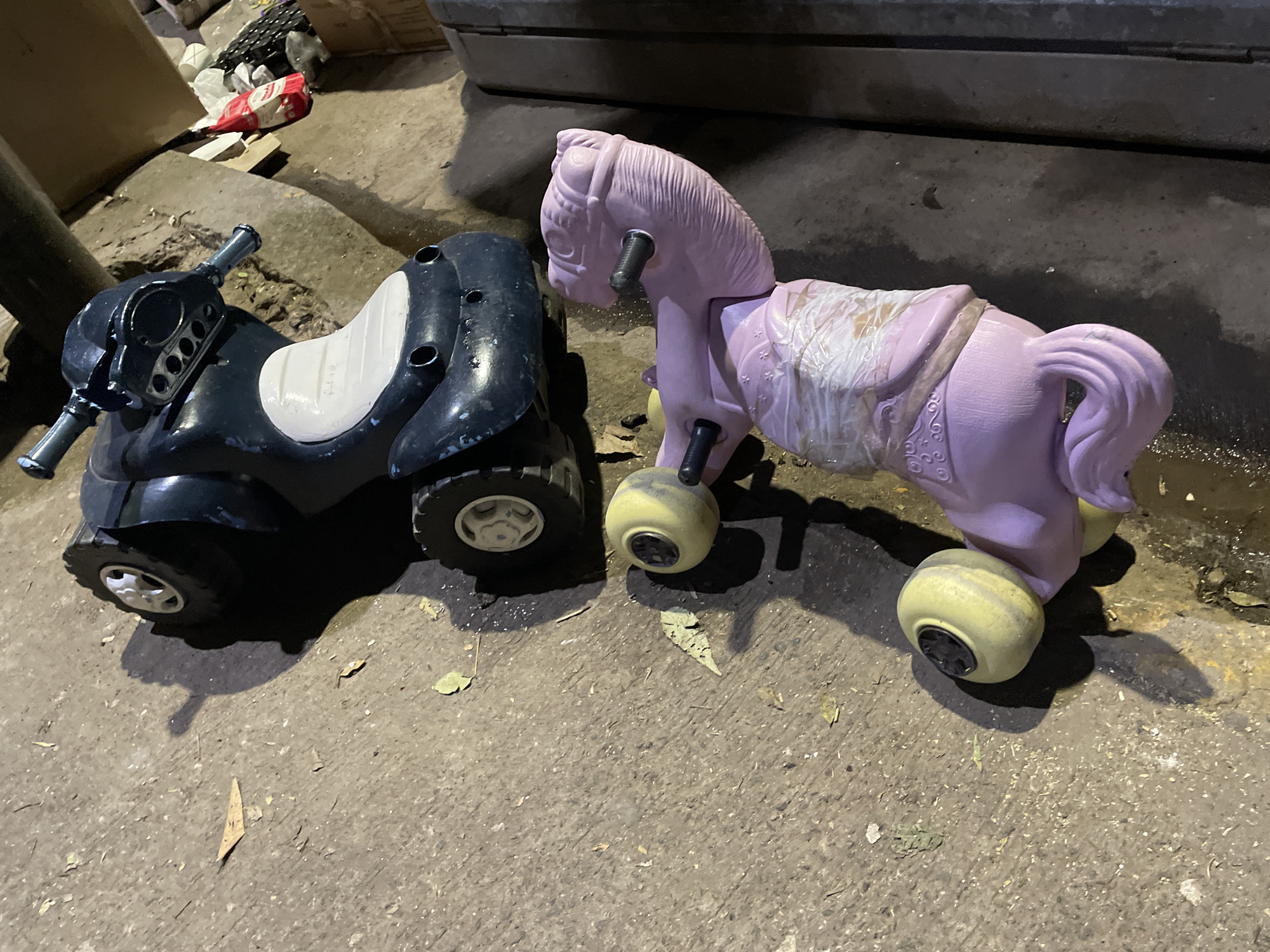 The image depicts two children's ride-on toys on a concrete surface. On the left is a black four-wheeled ATV-style vehicle with a white seat. To the right is a pink horse-shaped toy with four yellow wheels. The toys appear to be outdoors in a location with dim lighting. In the background, there are various objects and what may be a wall or container. The location in Buenos Aires, Argentina, cannot be determined from this image alone.