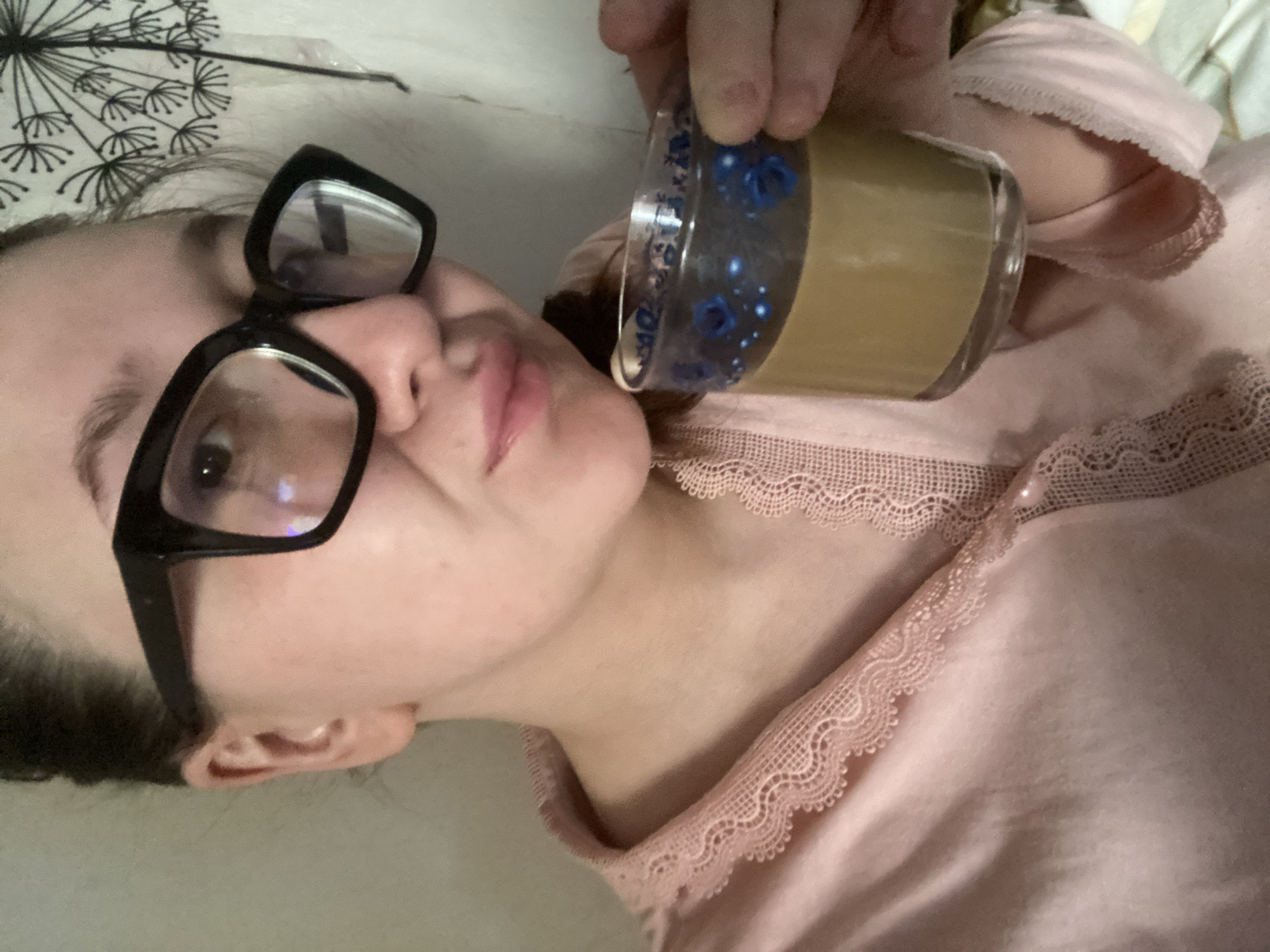 A person, likely female, is depicted in a close-up, indoor selfie, presumably taken in Chaikovsky, Russia. The individual wears a light pink, lace-trimmed pajama top and large black-framed eyeglasses. They hold a clear glass cup, decorated with a blue floral pattern, containing a light brown beverage in their right hand, positioned near their mouth. The background consists of a plain light-colored wall with a black dandelion silhouette sticker or drawing on the upper right.