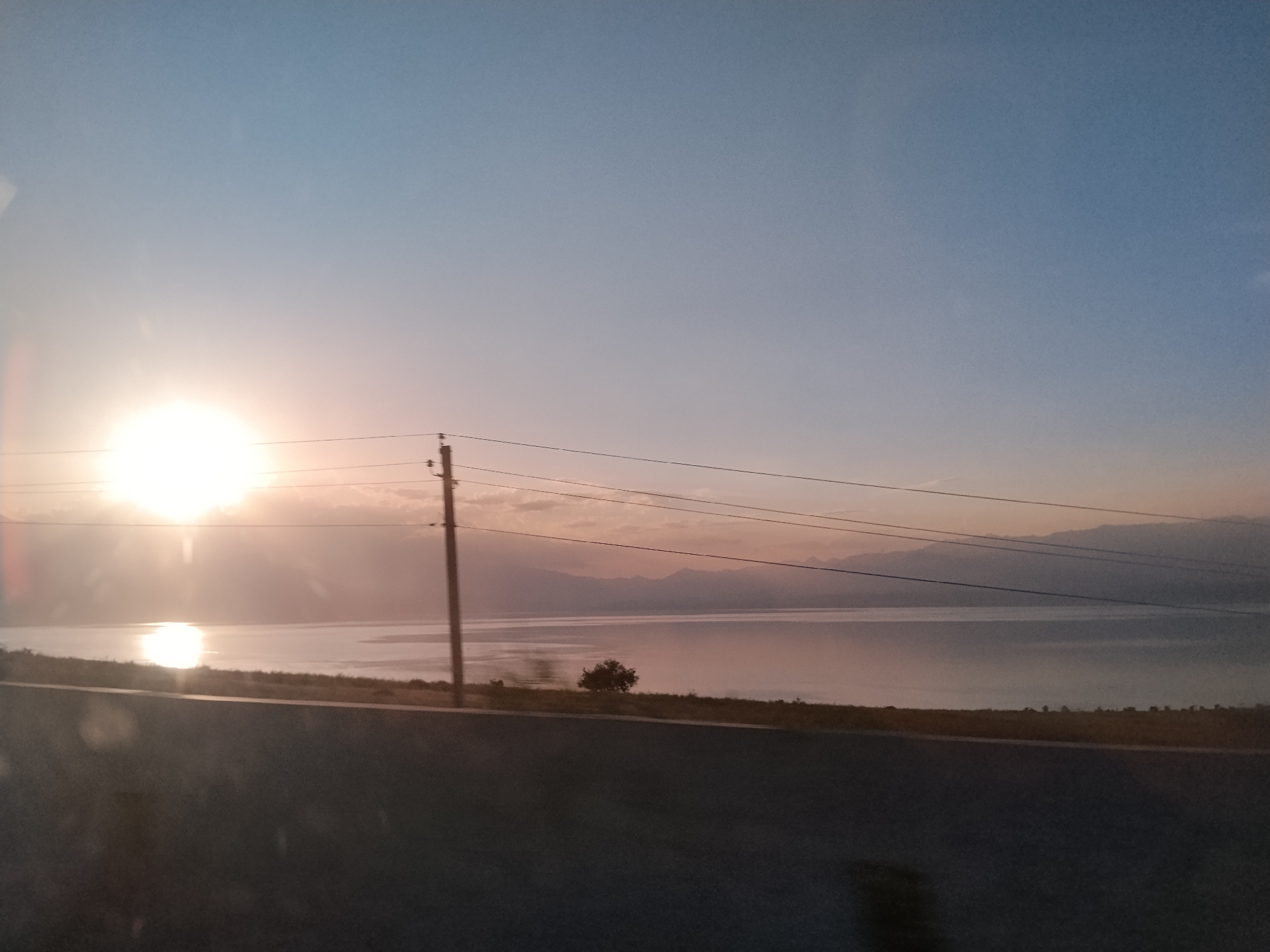 The image captures a serene sunset over a body of water, likely a lake or sea, with mountains visible in the distance. The sky transitions from a pale blue at the top to hues of orange and pink near the horizon, illuminated by the setting sun. The sun itself is a bright, glowing orb positioned on the left side of the frame, casting a shimmering reflection on the water.
In the foreground, a dark, paved road runs horizontally across the bottom of the image, suggesting the photograph was taken from a moving vehicle. To the left of the road, a grassy embankment with sparse vegetation and a solitary tree is visible. A utility pole with several power lines stretches across the middle ground, connecting to a pylon on the right. The overall impression is one of tranquility and natural beauty, characteristic of a rural or semi-urban landscape at dusk. The specific location in Chkalov, Kyrgyzstan, is not visually evident from the image alone, but the presence of mountains and a large body of water are consistent with geographical features found in that region.