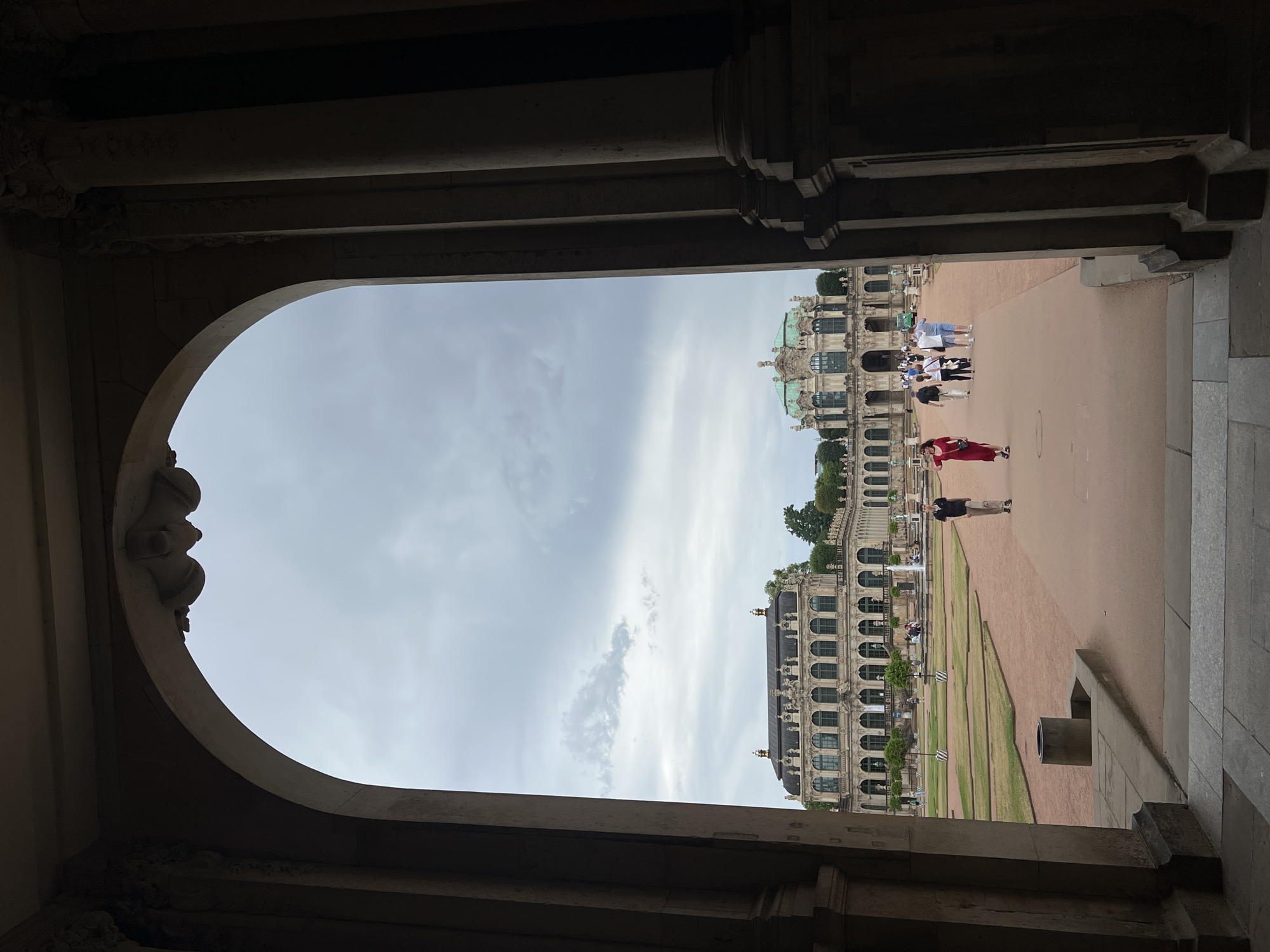 The image shows a view of the Zwinger complex in Dresden, Germany, seen through a large arched passageway. The Zwinger is a baroque palace and garden complex in Dresden, built by Matthäus Daniel Pöppelmann. In the foreground, the dark shadows of the archway frame a view of the Zwinger's courtyard and the buildings beyond. The courtyard has a paved walkway and manicured gardens. Several people are visible walking in the courtyard. On the left, a building with a long, multi-story facade featuring numerous arched windows is visible. On the right, a more ornate building with a green domed roof is prominent. The sky above is overcast.
