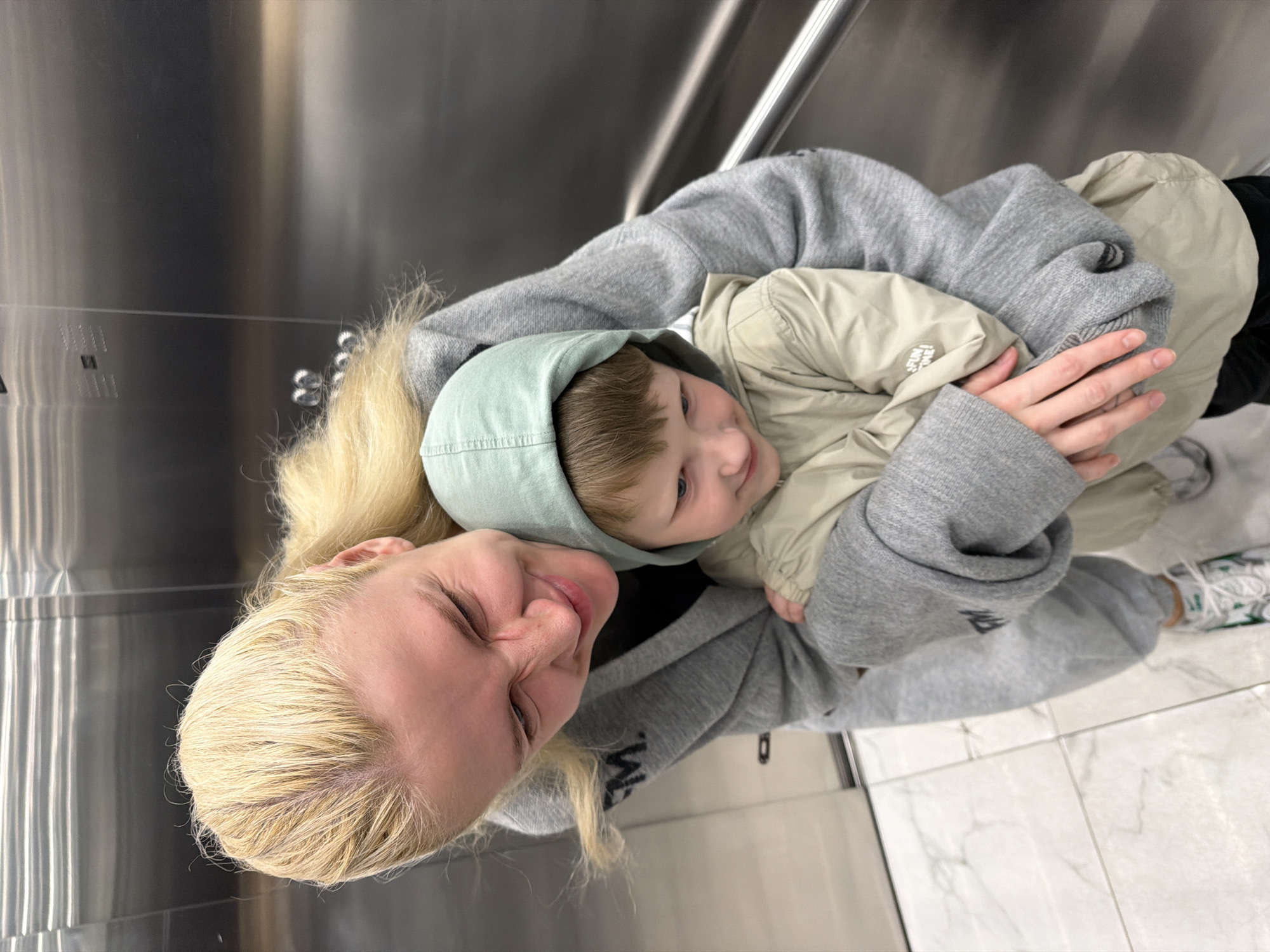 A woman with bleached blonde hair is holding a small child in her arms. The child is wearing a light green hooded jacket and a tan puffer jacket. The woman is wearing a gray hooded sweatshirt. The background appears to be an elevator interior with stainless steel walls. The floor appears to be light colored tile. The location is inferred to be in Belgrade, Serbia, but no explicit evidence exists in the image.