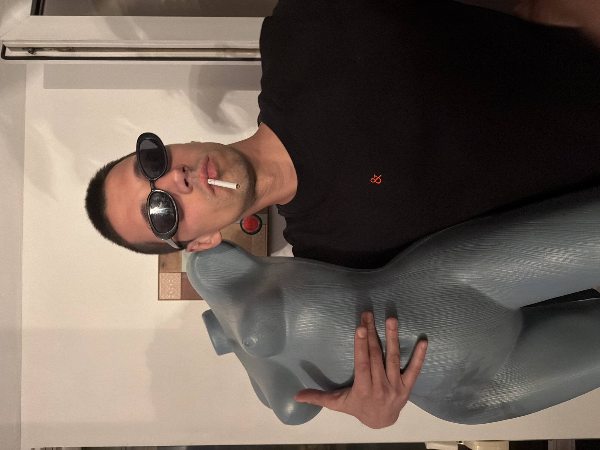 A man is depicted indoors in Sofia, Bulgaria, posing with a grey-blue female torso mannequin. The man, positioned on the right side of the frame with his head tilted, wears dark, oval-shaped sunglasses and a black short-sleeved t-shirt. On the left chest of his shirt, there is a small orange logo resembling a stylized infinity symbol or two interconnected loops. A cigarette is held between his lips. His hair is short and dark.
He has his left arm wrapped around the mannequin, which has a textured surface with horizontal lines and lacks a head or arms. The background consists primarily of a plain light-colored wall. In the upper left corner, a portion of a white window frame is visible. Behind the man's head, a small decorative wall accent with square, tiled patterns in various light brown and beige hues can be seen. The indoor lighting appears bright and even. The man's expression is partially obscured by his sunglasses, but his pose with the cigarette and the mannequin suggests a casual, possibly cool demeanor.