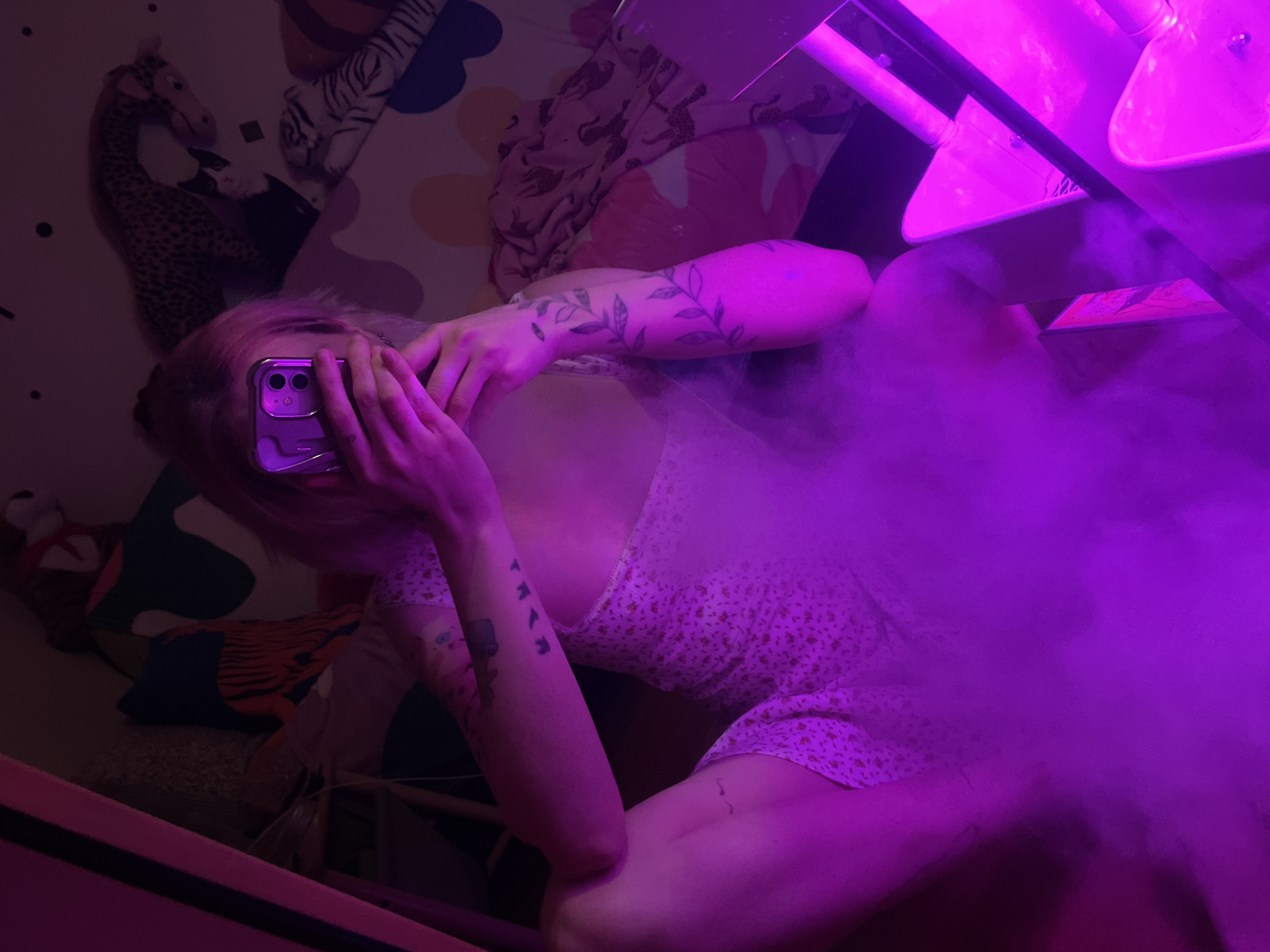 The image depicts a person in an indoor setting, likely a bedroom in Belgrade, Serbia, bathed in a strong purple light that creates a mysterious, ethereal atmosphere. Dense smoke or haze permeates the right side of the frame, generated by an object in the upper right corner that emits the vivid purple illumination.
The main subject is a person, likely female, with light-colored hair, reclining or lying down. Their face is obscured by a purple smartphone (an iPhone with dual cameras in a translucent purple case) held directly in front of it. They are wearing a light-colored dress with a small, floral pattern. Visible on their arms are multiple tattoos; one arm displays a botanical design of leaves or vines, while the other features smaller, abstract symbols.
In the background, behind the person, bedding or a blanket with various patterns is visible, including stuffed animals like a giraffe and a tiger, suggesting a personal living space. The image is taken from a high-angle perspective. No text is visible.