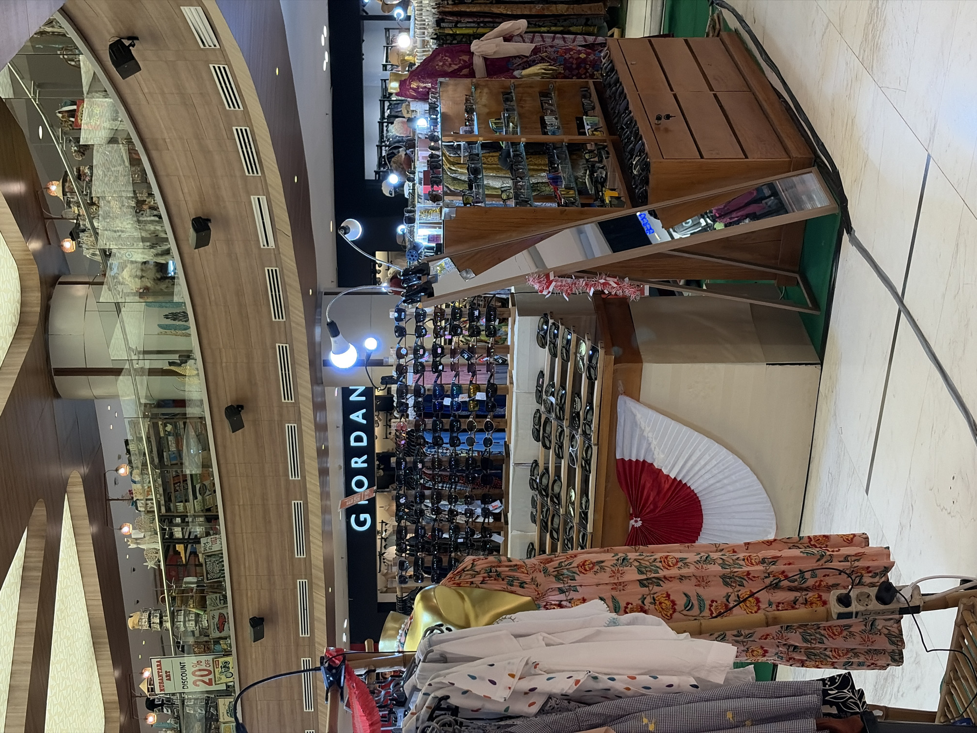 The image captures a section of a multi-level indoor shopping mall in Kuta, Indonesia. In the foreground, clothing racks display various garments, including a patterned dress on a mannequin. To the right, a prominent display of sunglasses is arranged on shelves and in drawers. Behind the sunglasses, a store with the signage "GIORDAN" is visible, featuring racks of apparel. The mall's architecture is characterized by curved wooden panels and tiered levels with railings. Lighting fixtures are integrated into the ceiling and displays. The floor is tiled in a light color with subtle patterns. There are no discernible people actively engaged in shopping or transactions within the frame.