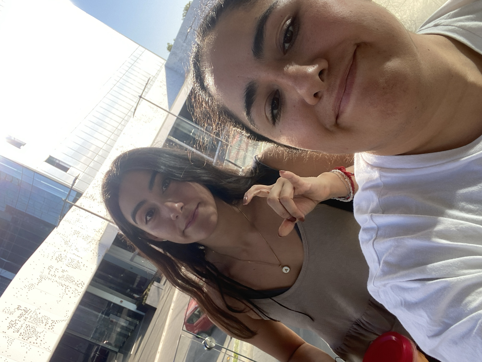 Two women are in the foreground of a photograph, with a building in the background. The photograph is taken at an upward angle. The woman on the right is wearing a white shirt and has dark hair pulled back. The woman on the left is wearing a beige tank top and has long, dark hair. She is wearing a necklace with a round pendant. She is holding up her hand with her index finger extended. In the background, there is a modern building with glass and metal elements. The building is located in Puebla, Mexico.