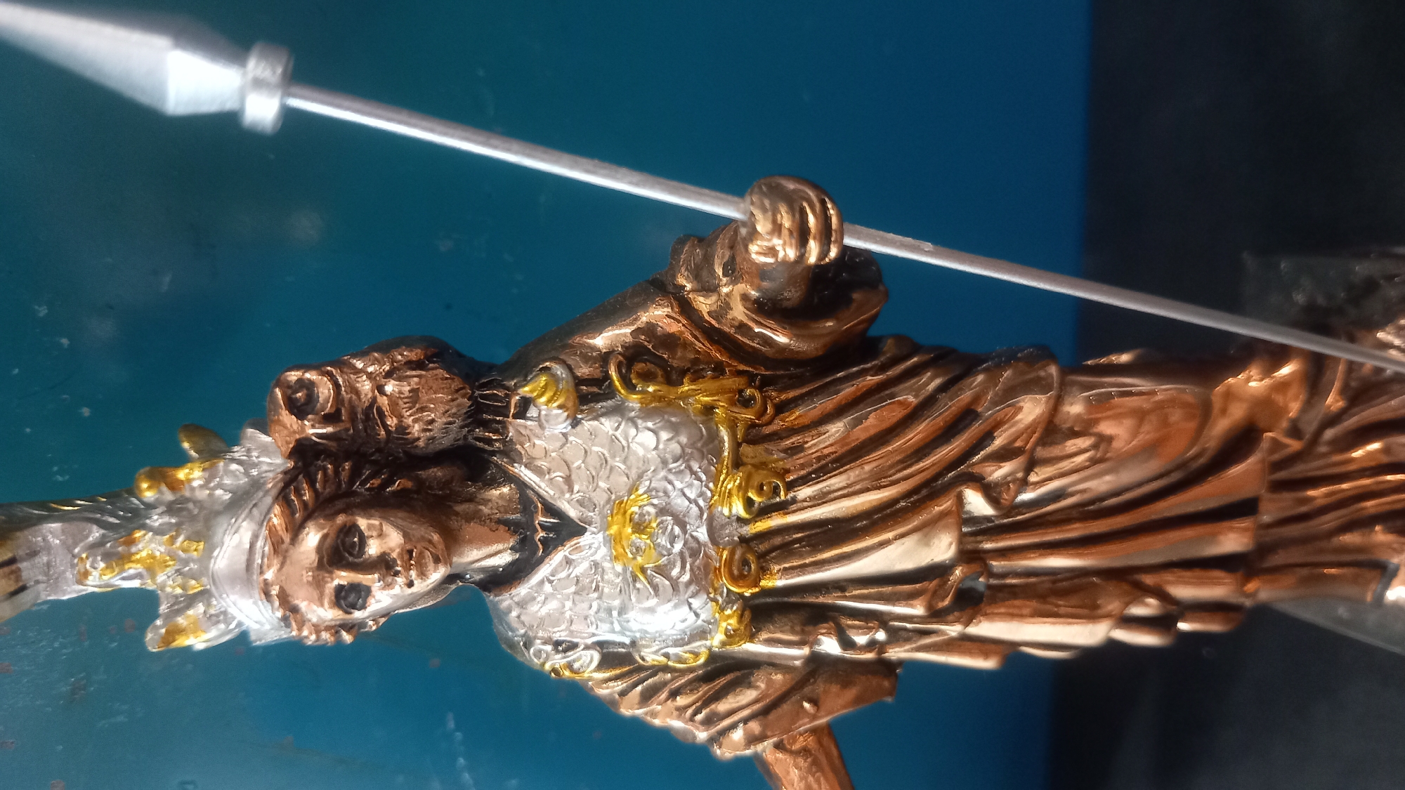 The image shows a close-up, slightly angled shot of a bronze statue of Athena, the Greek goddess of wisdom and warfare. The statue depicts Athena wearing a helmet and ornate armor, with a spear held in her right hand and an owl perched on her left shoulder. The statue's metallic sheen is evident, with gold accents on her armor and helmet. The background is a blurred, solid blue surface, likely a wall or backdrop. There is no identifiable indication of location in the city of Kos, Greece, within the image itself.