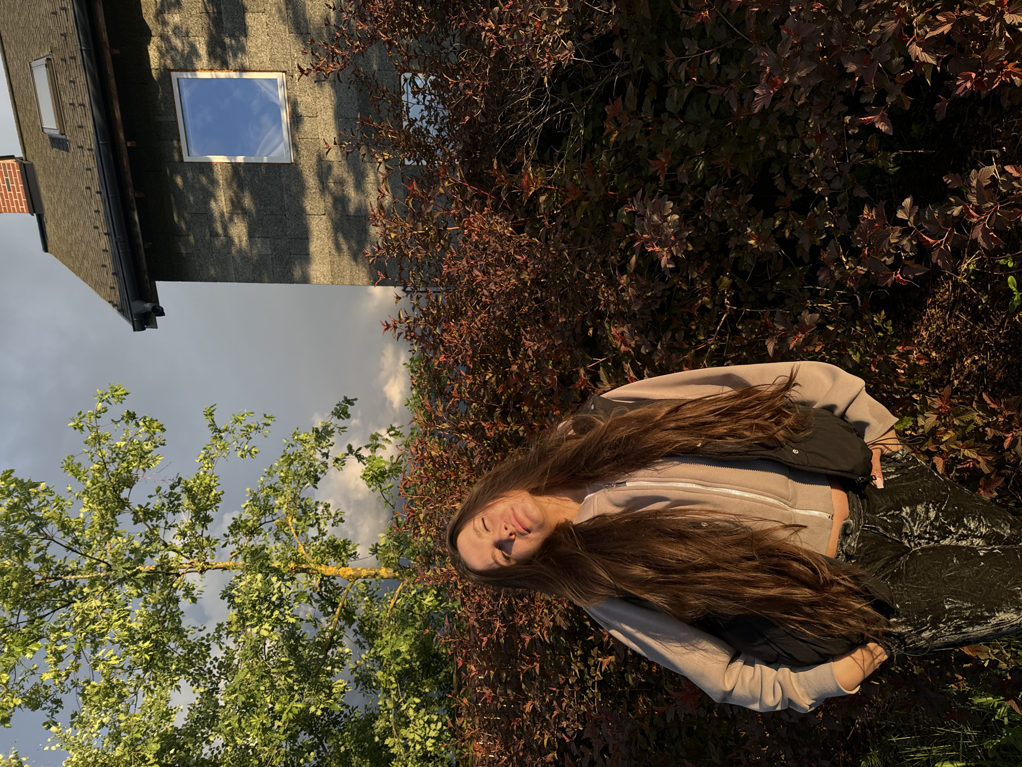 The image shows a young woman with long brown hair standing in a garden or park-like setting. She is wearing a light-colored zip-up jacket and dark-colored pants. Behind her, there is a mix of green trees and bushes with reddish-brown foliage. To the left, a corner of a building with a window is visible against a cloudy sky. The location is not explicitly stated within the image, and therefore it is impossible to verify if it is located in the city of Vysokinichi, Russia.