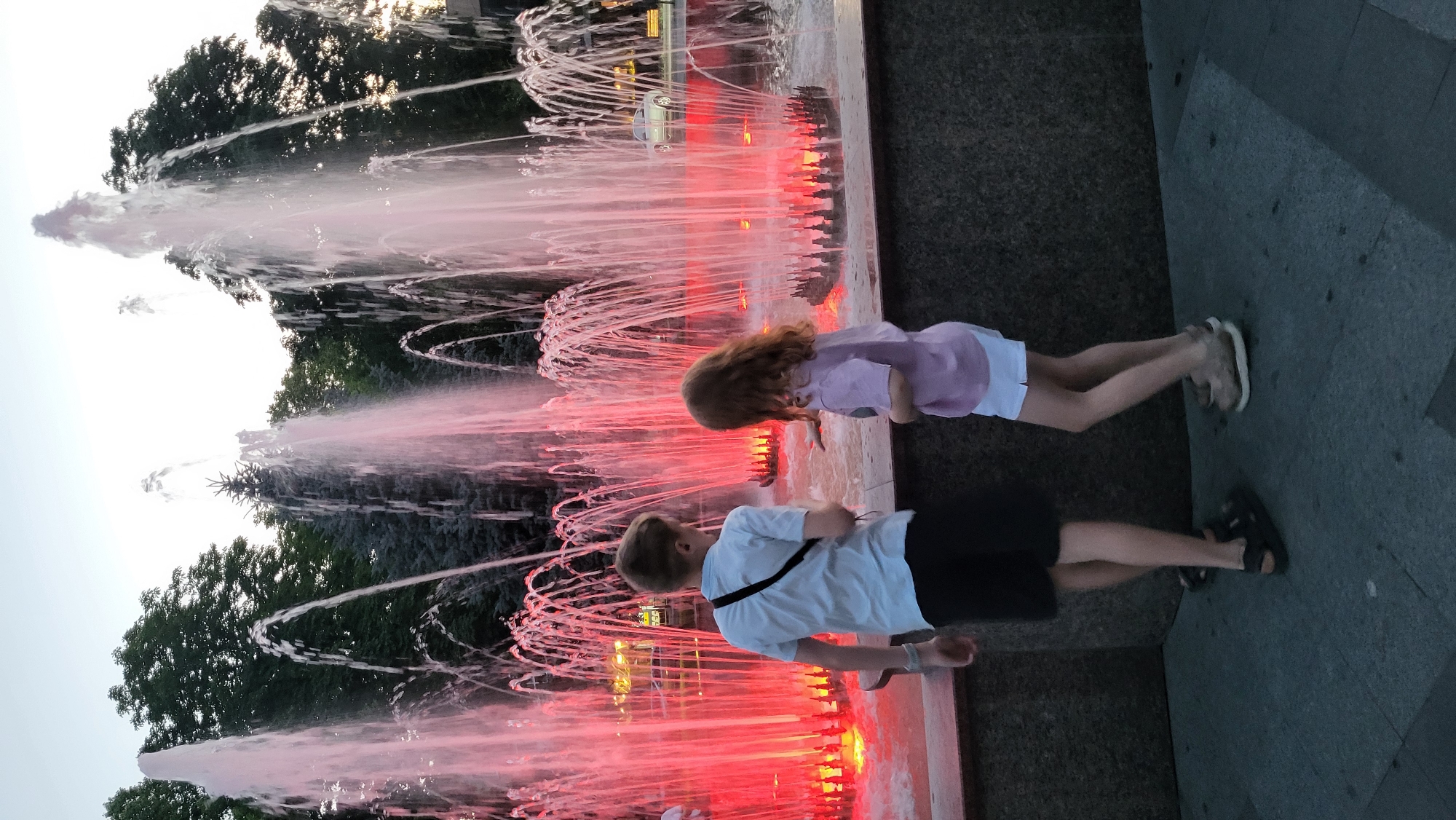 In an outdoor urban setting, two children, a boy and a girl, stand near a fountain illuminated with red lights. The boy, facing away from the camera, wears a light blue short-sleeved shirt and dark shorts, with a black strap across his chest and black sandals. The girl, also facing away, has reddish-brown hair and is dressed in a light purple t-shirt and white shorts, wearing light-colored sneakers. They are positioned on a paved surface in front of a stone structure that forms the base of the fountain. The fountain itself features numerous streams of water cascading downwards, with bright red lights creating a dramatic effect. In the background, dark green trees and a bright sky are visible, along with what appears to be a light-colored car parked in the distance. The lighting suggests it is dusk or early evening.