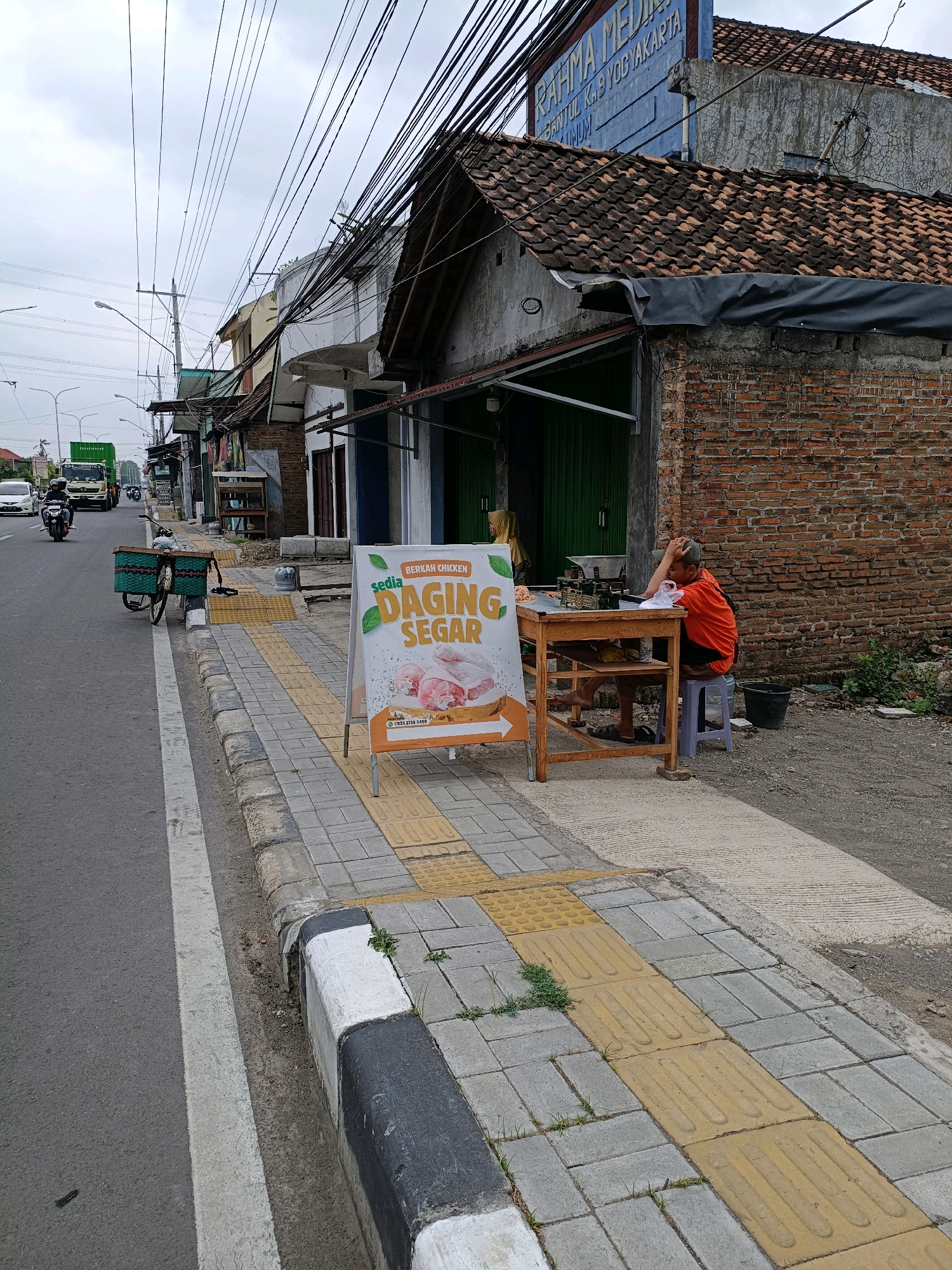 The image depicts a street scene in Sewon, Indonesia. A busy road runs parallel to a sidewalk paved with grey and yellow textured tiles, indicating a pedestrian path. Along the sidewalk, a white A-frame sign advertises "BERKAH CHICKEN" with an image of raw chicken parts. To the right of the sign, a man in an orange shirt sits at a wooden table, resting his head in his hand. A bicycle with a green basket is parked on the sidewalk near the sign. In the background, buildings with tiled roofs line the street, and power lines crisscross overhead. A white car and a green truck are visible on the road, along with a motorcycle. The overall environment suggests an urban commercial area.