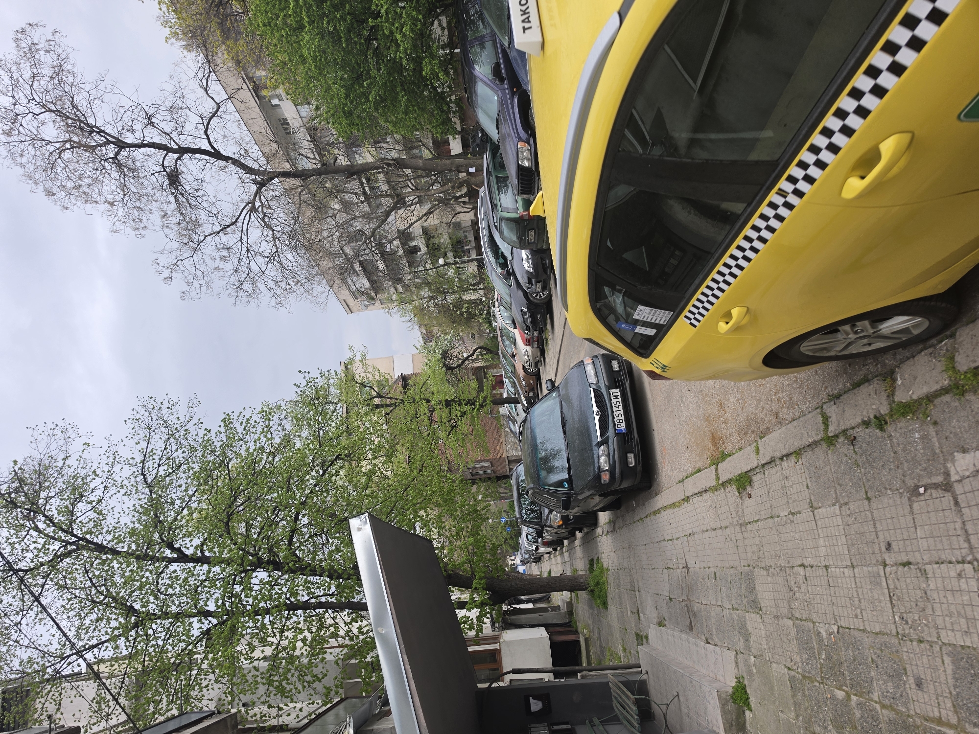 The image captures a street scene in Plovdiv, Bulgaria. The perspective is from the sidewalk, looking along a street lined with parked cars. A yellow taxi with checkered markings is prominently featured on the right side of the frame. Parked behind the taxi are several other vehicles, mostly dark-colored. Buildings are visible in the background, partially obscured by trees with budding leaves. The sky is overcast. A sign or awning structure is visible on the lower left, extending partially into the frame. The sidewalk is paved with square stones. A license plate of a car parked alongside the curb reads "PB 5145 MT".