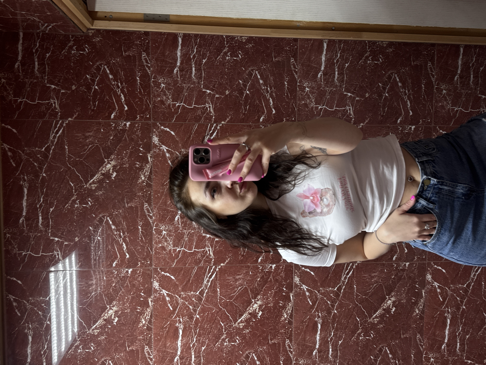 A young woman with dark, wavy hair takes a mirror selfie. She is wearing a white t-shirt with a graphic of a pink flower and is holding a pink phone in her right hand. Her left hand is in the pocket of her blue denim jeans. She has a silver bracelet on her left wrist and a ring on her right hand. Her fingernails are painted bright pink, and she has a belly button piercing. The background is a wall with red marble tiles and a wooden door frame on the right side. A light fixture is visible in the reflection on the ceiling. The scene appears to be indoors, possibly in a bathroom or dressing area.