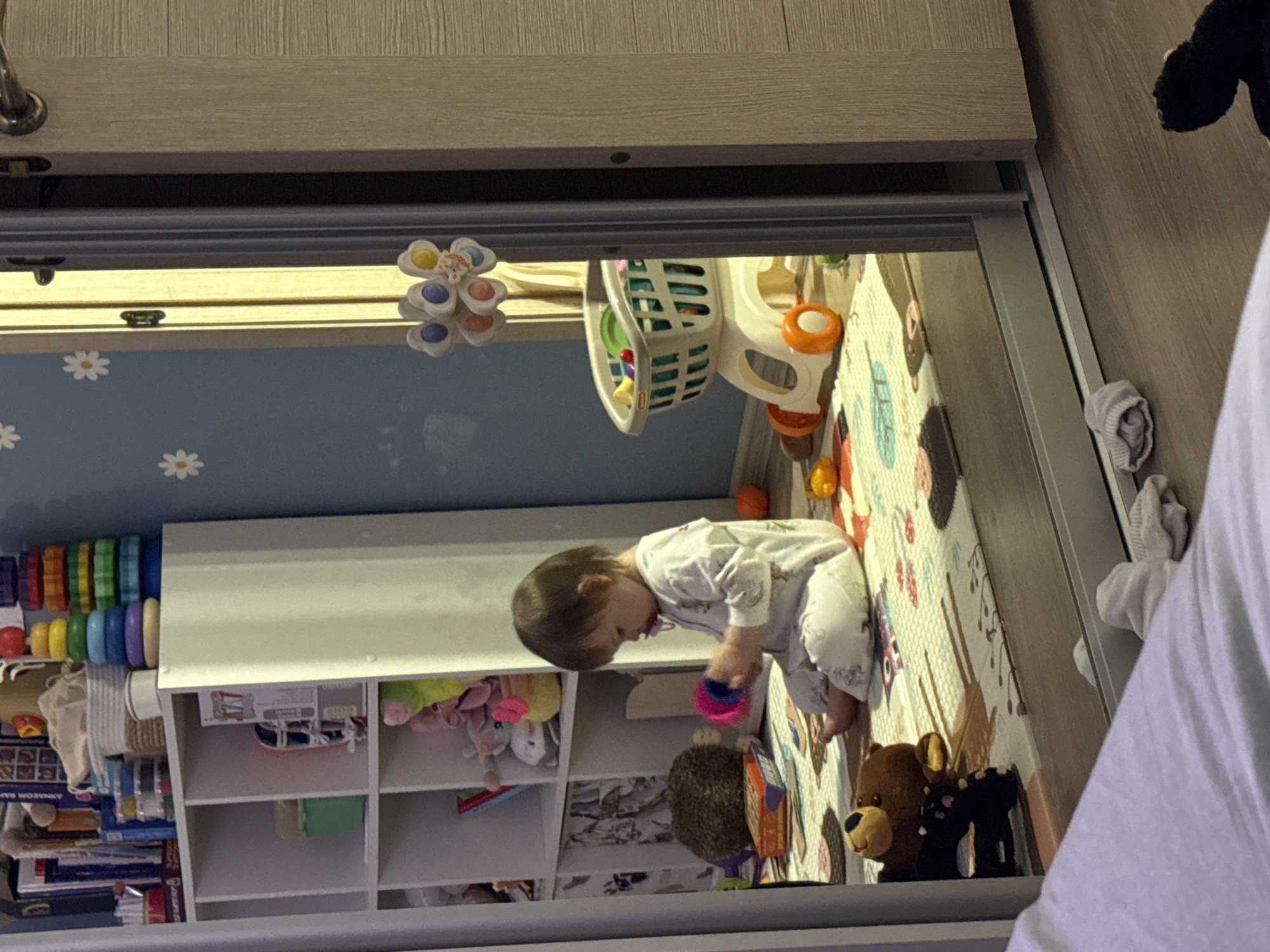A child in pajamas is playing with toys on a mat in a room. The room contains shelves with books and toys. A toy shopping cart is also visible. The location and city of Moscow, Russia, cannot be determined from the image.