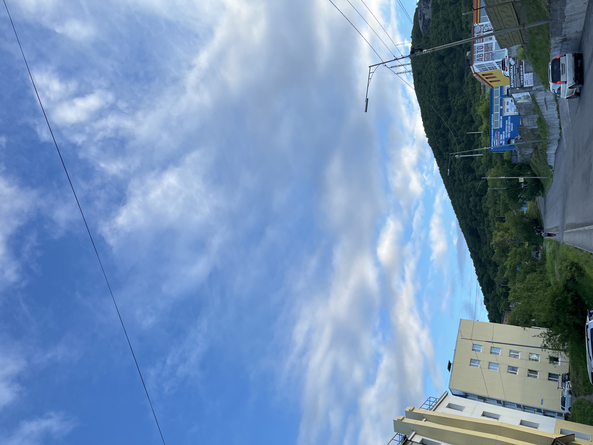 This image captures an upward view of a street in Petropavlovsk-Kamchatsky, Russia, with a prominent hill covered in dense green trees dominating the background. The sky is a vibrant blue, streaked with wispy white clouds, and a thin black wire stretches diagonally across it. On the left side of the street, a tall, pale yellow apartment building with multiple windows is visible. Further up the street, on the right, are several commercial buildings, including one with large signs advertising "ABTO" and "ABTOСЕРВИС" (AUTO and AUTO SERVICE). A white SUV is parked on the right side of the road, and another vehicle is visible further down. A few individuals can be seen walking up the street. The overall scene depicts a somewhat hilly urban landscape with a mix of residential and commercial structures, set against a backdrop of natural greenery and a dynamic sky.