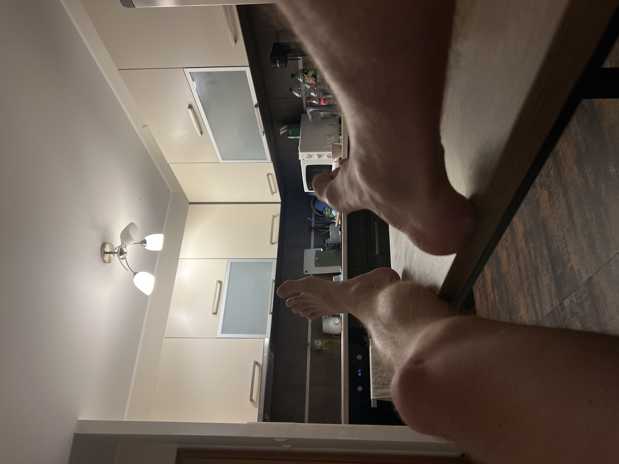 The image shows a first-person perspective of a person's bare feet resting on a table in a kitchen. The kitchen has cream-colored cabinets, dark countertops, and a tiled floor. There are various kitchen appliances visible, including a microwave, a coffee maker, and a refrigerator. The lighting suggests it is nighttime or indoors with artificial light. There is no specific indication of the location being in Rumia, Poland; this detail is an external assertion.