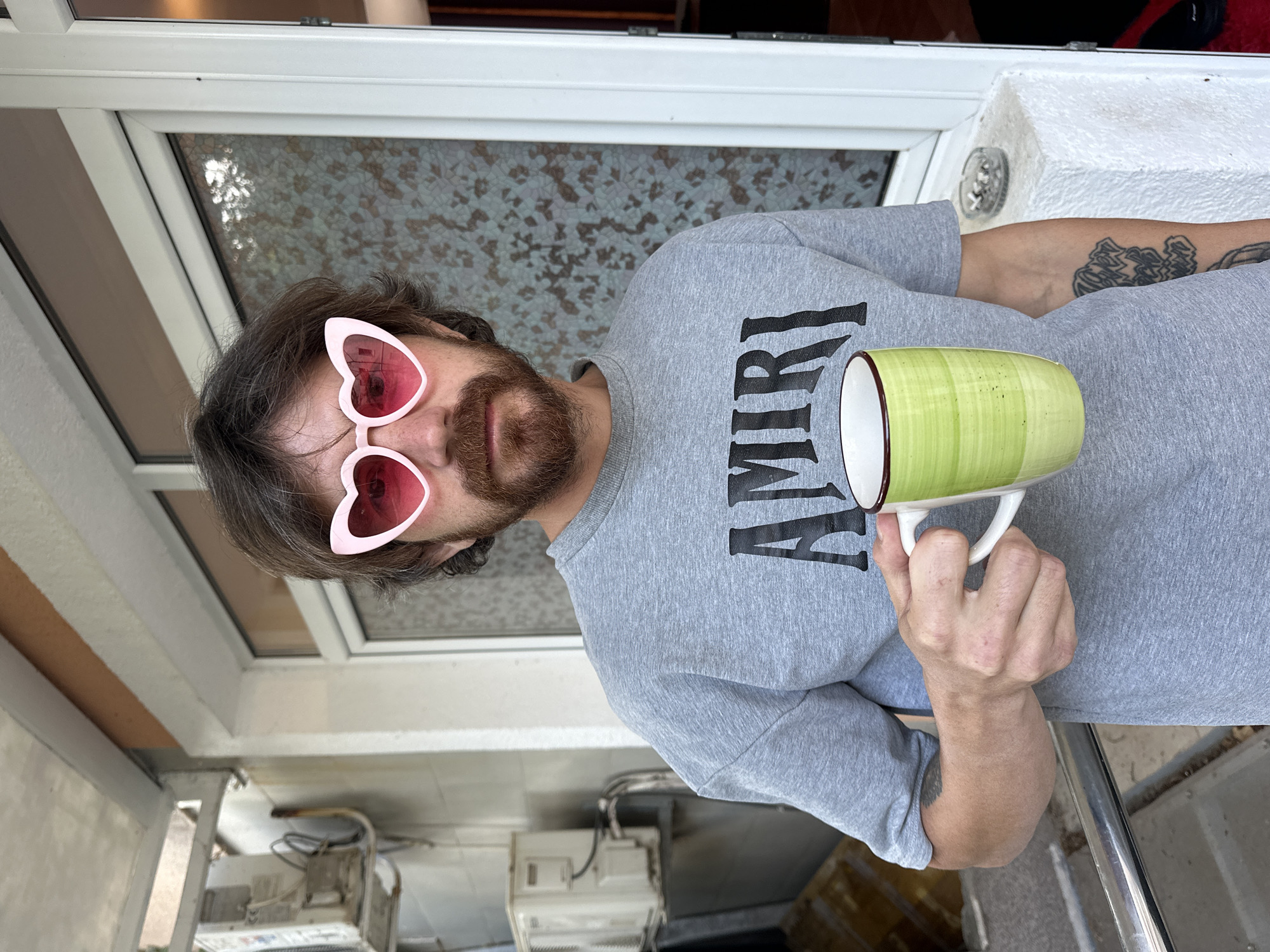 A man with a beard and mustache wearing pink heart-shaped sunglasses and a grey "AMIRI" t-shirt holds a green and white striped mug. He stands indoors, near a window with textured glass. A tattoo is visible on his left forearm. Other objects, including a washing machine, can be seen in the background. Without additional information, it's impossible to confirm the location in Budva, Montenegro.