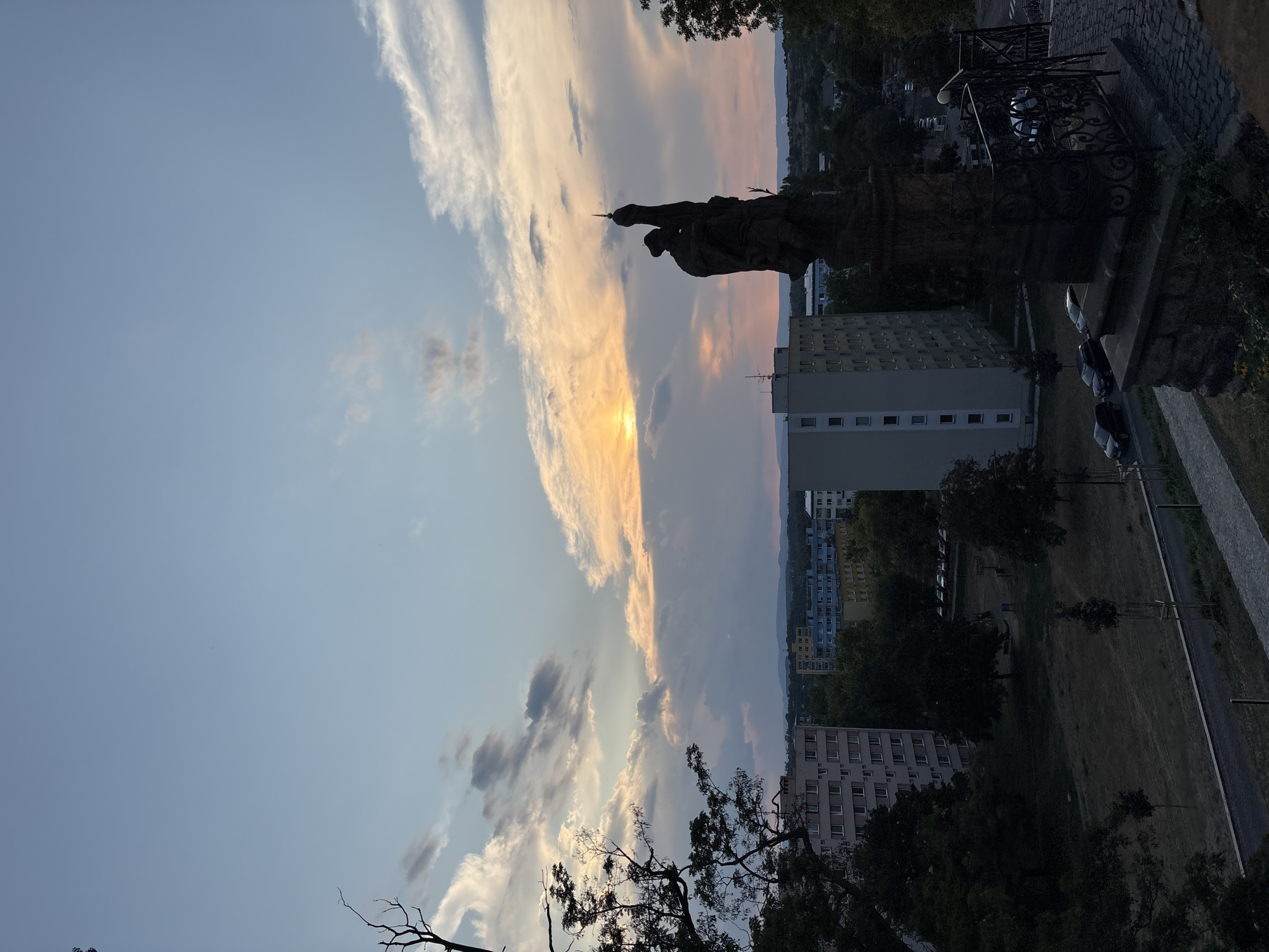 A statue of a robed figure holding a pointed object stands in silhouette against a dramatic sunset sky. The statue is dark brown and weathered, with intricate details visible on its surface. It is mounted on a stone pedestal, surrounded by a black wrought-iron fence with decorative scrollwork. In the background, a city skyline is visible, with several tall, apartment buildings and trees. The sky is a mix of blue, orange, and pink, with clouds scattered throughout. The sun is setting on the left side of the image, casting a warm glow on the clouds. The scene appears to be set on an elevated vantage point, overlooking the city. The overall impression is one of twilight in an urban setting, with a historical monument as the central focus.