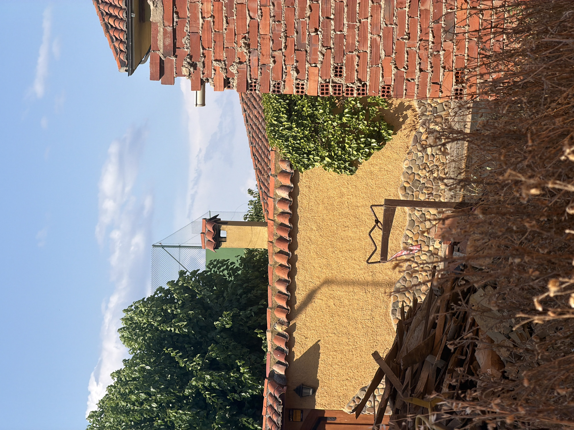 The image depicts an outdoor scene in Arcahueja, Spain, on a clear day with a blue sky dotted with wispy clouds. The foreground is dominated by dry, brown vegetation. In the midground, a textured yellow wall forms a prominent feature, topped by a terracotta tiled roof. A section of a red brick wall, partially constructed and with visible hollow bricks, stands to the right, partially obscuring the view of the yellow wall and roof. A chimney with a terracotta cap rises above the yellow wall. To the left of the yellow wall, a dark wooden door is visible. A pile of discarded wooden planks and rubble sits in front of the yellow and stone-patterned lower wall. Green ivy climbs on the yellow wall near the brick structure. In the background, a lush green tree with dense foliage is visible, and a chain-link fence with a green surface behind it suggests a recreational area, possibly a tennis court. A metal pole with a curved top extends from the rubble pile, and a small red and white flag is attached to it. A small light fixture is mounted on the yellow wall. The overall impression is one of a partially maintained or under-construction property.