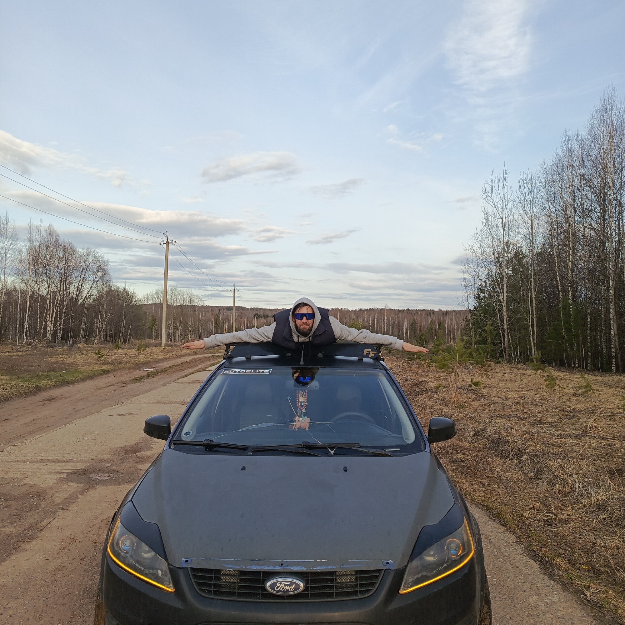 A man lies prone on the roof of a dark grey Ford Focus car, arms outstretched, in a rural setting near Марковский, Russia. He wears a grey hooded sweatshirt, a black vest, and sunglasses. The car has an "AUTODELITE" branded roof rack, a Ford logo on the grill, and a cracked windshield with a hanging ornament. The background includes a dirt road, bare trees, a power line, and dry, grassy ground. The sky is lightly cloudy.