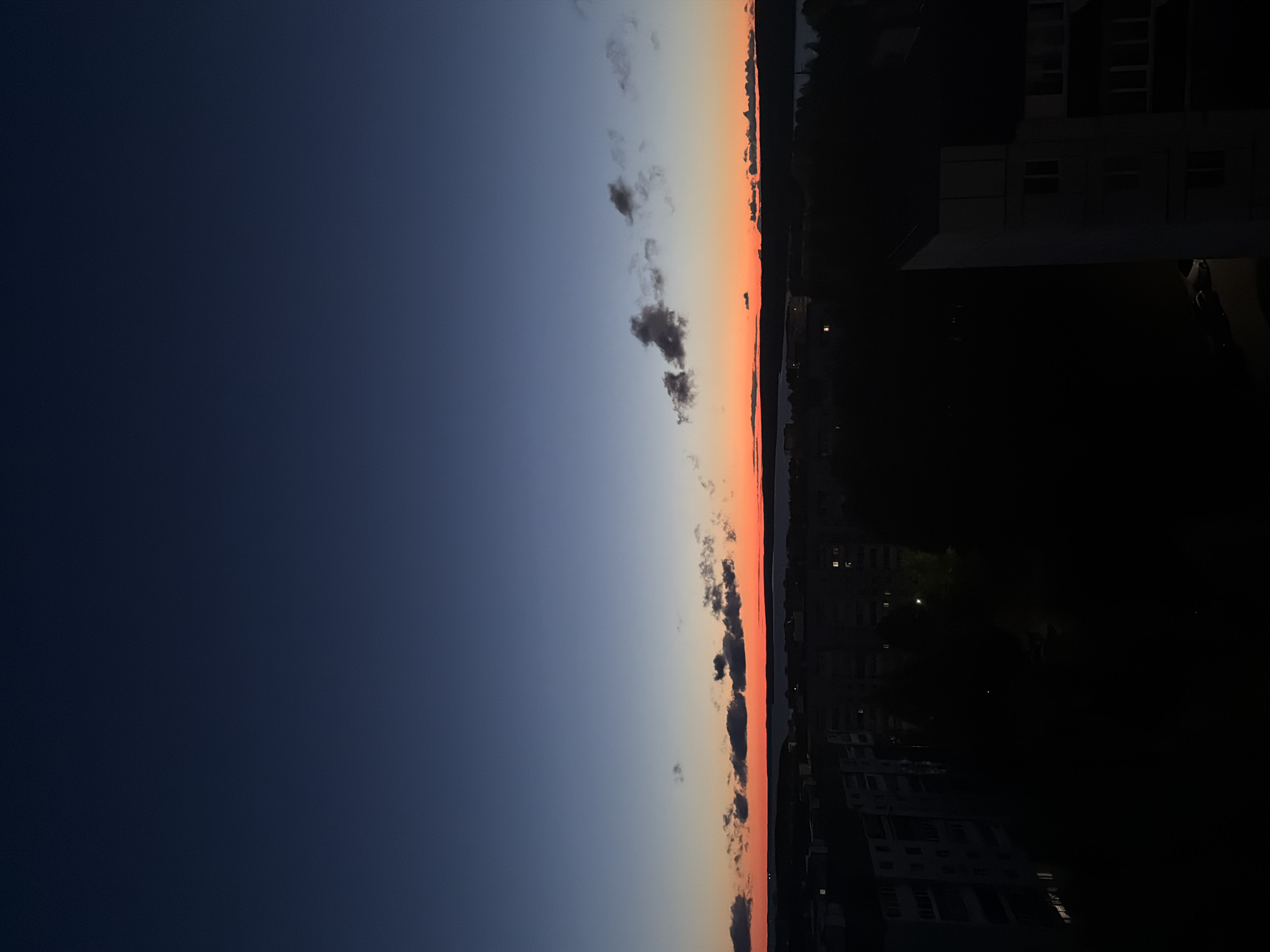 The image depicts a sunset over what appears to be a body of water near the city of Chaikovsky, Russia. The sky transitions from dark blue to a lighter blue, fading into orange and red hues near the horizon. Some scattered clouds are visible along the horizon line.
On the right side of the image, the cityscape is largely silhouetted and dark, indicating the setting sun is behind it. There are multiple high-rise buildings visible with some lit windows. The scene suggests a residential area situated near a river or lake, typical for a city like Chaikovsky.