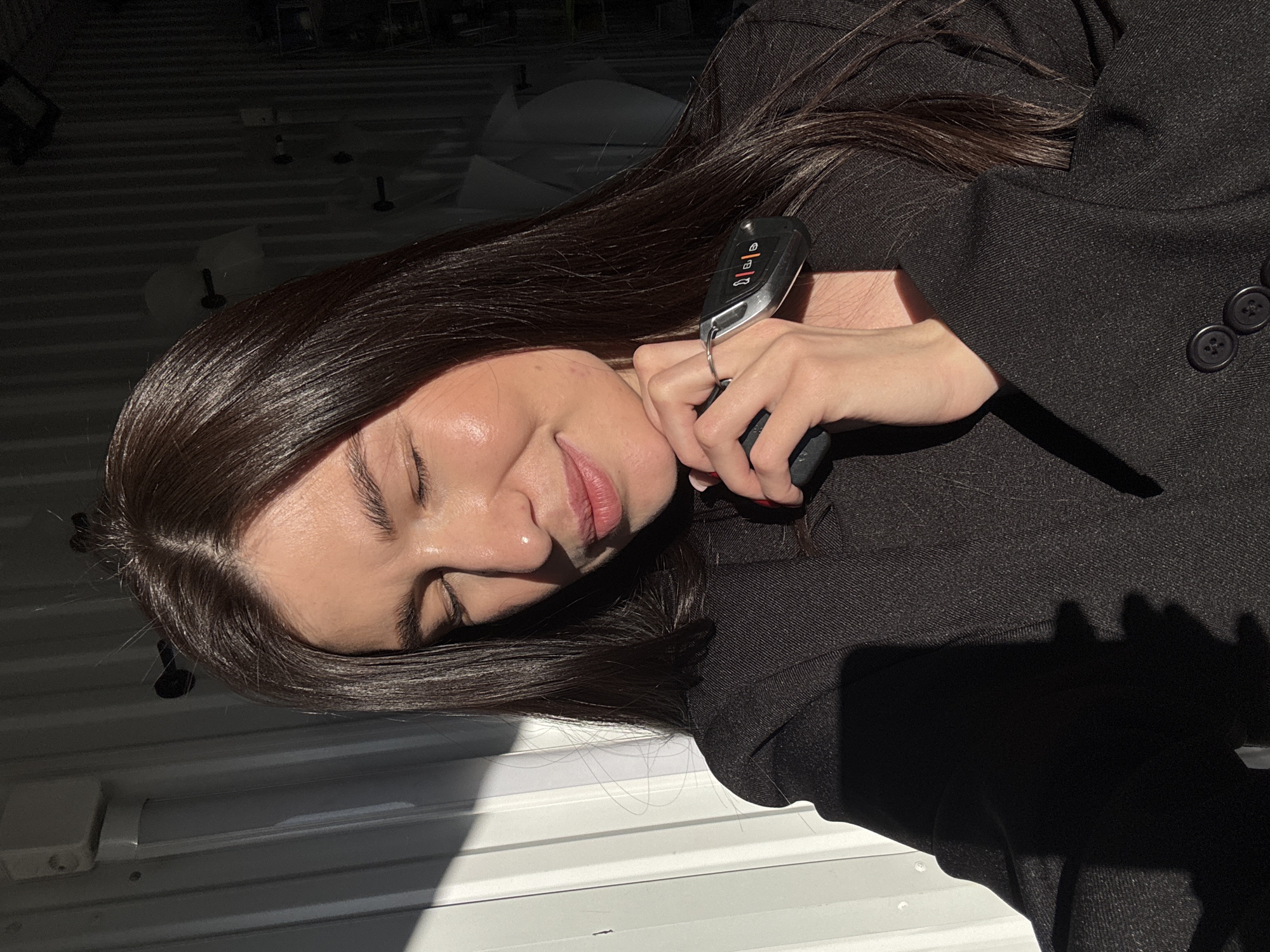 A woman with dark brown hair, wearing a dark blazer with two visible buttons, is lying on a white, horizontally striped surface. She is holding a car key in her right hand, which rests near her face. The location may be a garage in Chaikovsky, Russia. The background includes indistinct objects and shadows.