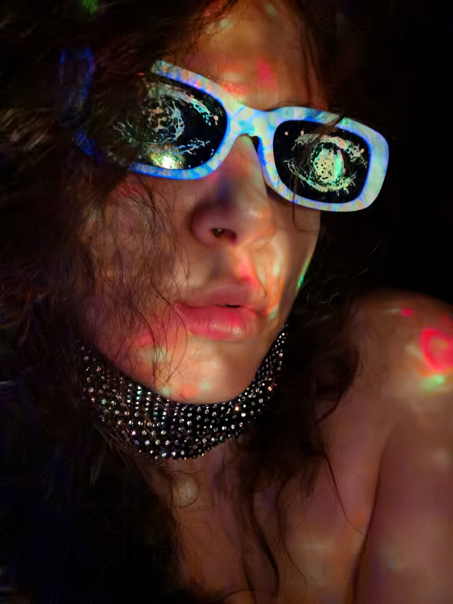 A close-up portrait of a person with curly dark hair wearing white-framed sunglasses with mirrored lenses. The lenses reflect a blurry scene with what appears to be light sources. The person is wearing a black choker with reflective, possibly rhinestone, details. The skin is illuminated with colorful light projections, mainly red, green, and pink. The background is dark, contributing to the disco or party-like atmosphere. There is no information available in the image to confirm the location is in Chaikovsky, Russia.