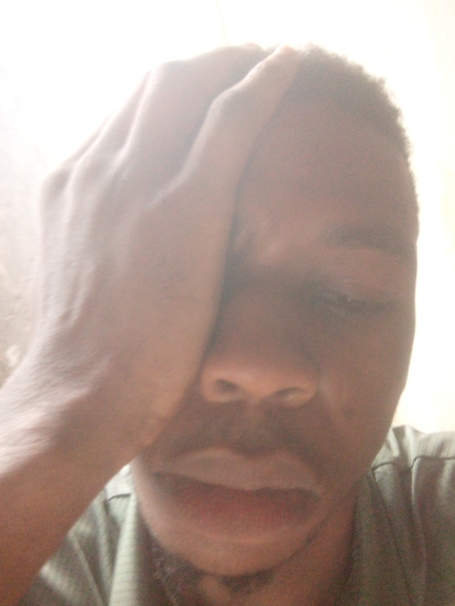 A close-up, poorly lit image of a man's face with his hand partially obscuring his forehead and eyes. His mouth is slightly open, and he has a mustache and short hair. He is wearing a shirt, but the color is unclear. The background is a blurry, light-colored wall. The location is Unknown, Unknown.