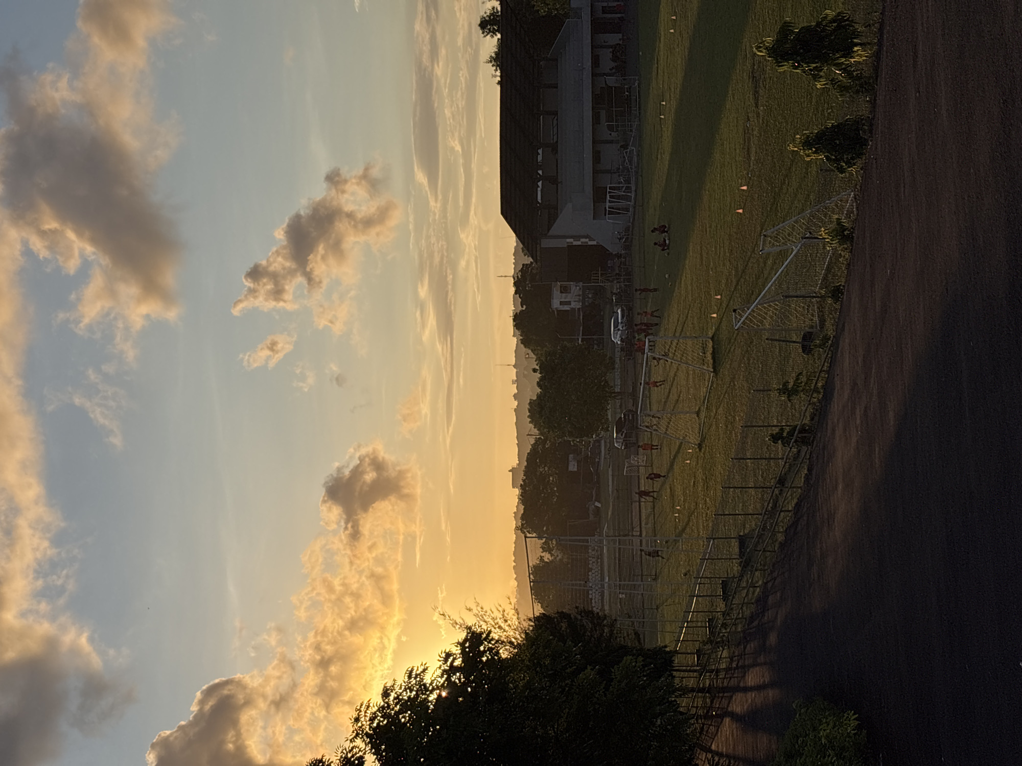 The image presents an outdoor scene at sunset. The sky is filled with clouds illuminated by the setting sun, casting a warm, golden light. On the right, a soccer field is visible, with soccer nets and cones set up. Individuals can be seen on the field, likely practicing. A building structure stands adjacent to the field. In the foreground, foliage frames the lower portion of the view. The location is unidentified but based on the prompt, it is likely located in Kuta Selatan, Indonesia.