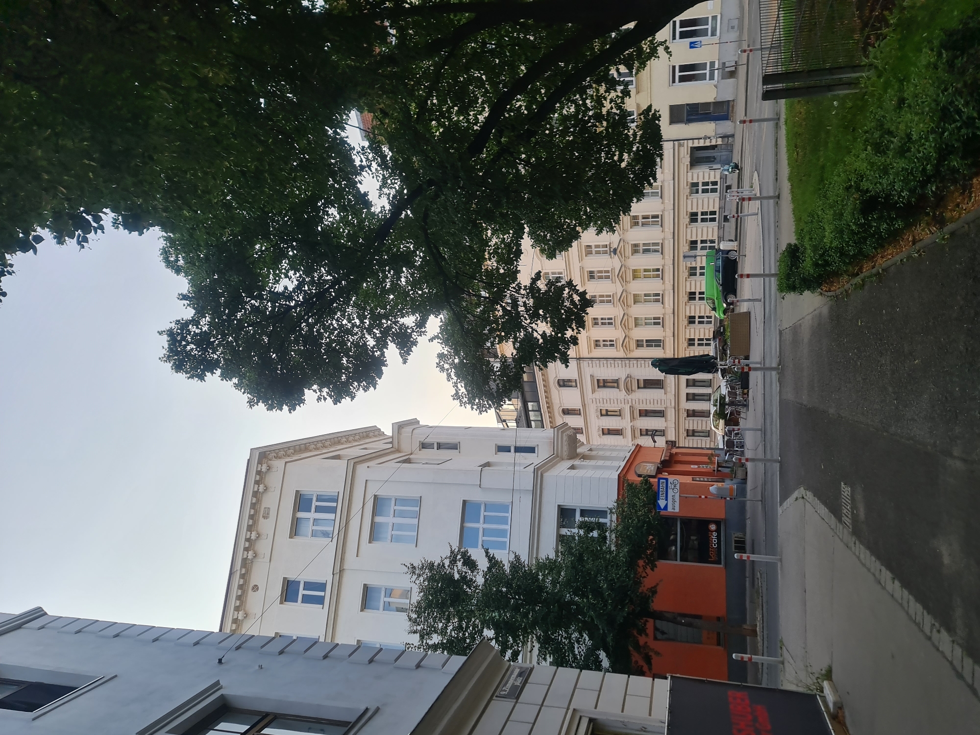 This is a street-level, eye-level, wide shot image of a street in Vienna, Austria, captured during the daytime. The scene features a prominent white building with multiple windows and ornate architectural details on the right side of the frame, and another white building on the left. A large tree with dark green foliage is positioned in the foreground on the right, partially obscuring the view of the buildings. In the middle ground, a street intersection is visible with several bollards and a sign indicating "Einbahn" (one-way). A bright green van and a dark colored car are parked on the street. Further down the street, an orange-fronted building with a sign that says "Lazareth cafe" is visible, with outdoor seating tables and chairs set up. The sky is overcast and light gray.