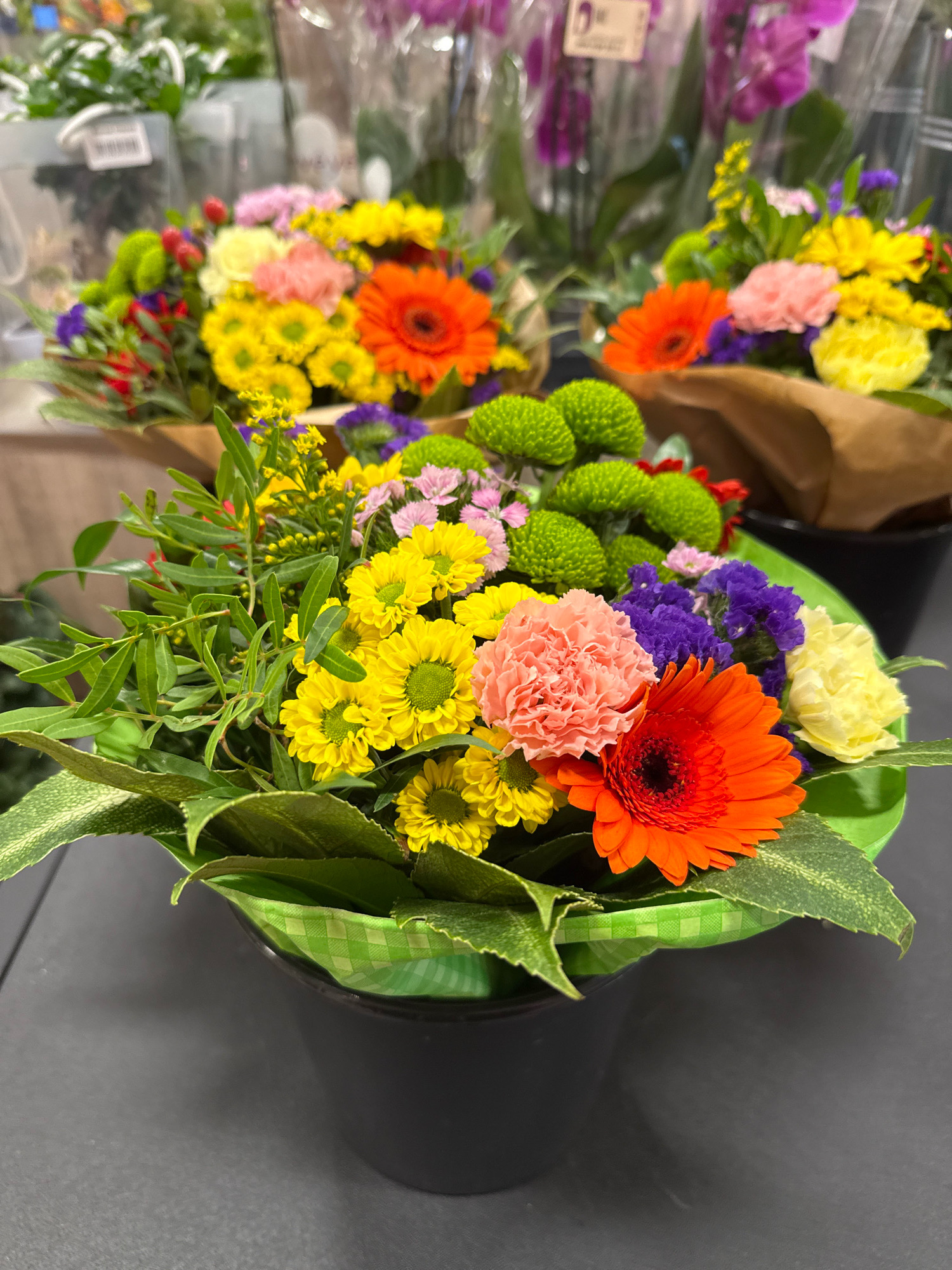 The image shows a close-up, eye-level view of a vibrantly colored bouquet of flowers presented in a dark gray cylindrical pot. The bouquet is composed of various flowers and foliage, including bright orange gerbera daisies, pink carnations, yellow chrysanthemums, small purple flowers, and green, leafy branches. The flowers are arranged in a loose, natural style, with some greenery spilling over the edges of the pot. A green ribbon with a white checkered pattern is visible around the rim of the pot. The background is slightly blurred, showing hints of other flowers and clear plastic packaging, suggesting the flowers are displayed for sale in a shop or market. The overall lighting is bright, highlighting the colors of the flowers. The scene is set against a dark gray, smooth surface, likely a tabletop or counter. This image was captured in Bad Lauterberg im Harz, Germany.