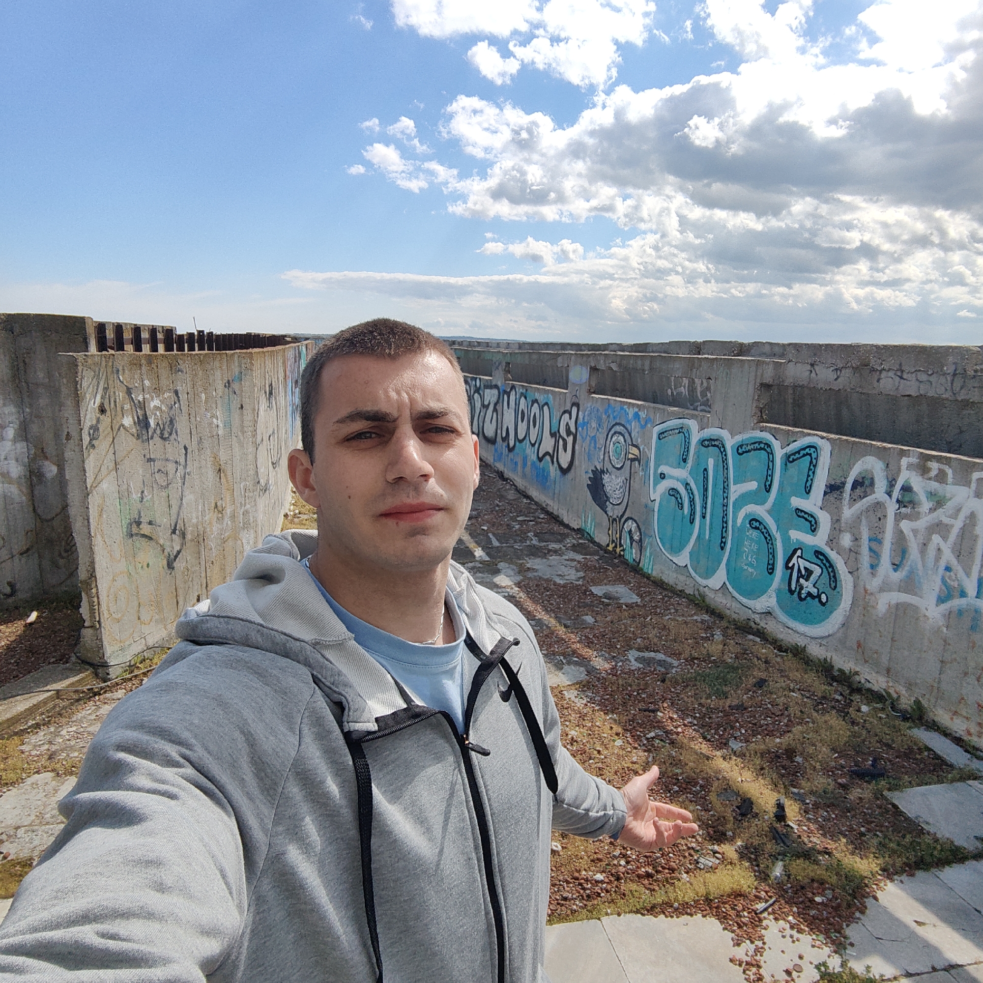 A man is taking a selfie outdoors. He is wearing a gray hoodie and a blue t-shirt. The background shows graffiti-covered concrete walls under a partly cloudy sky. The ground is covered with reddish-brown stones and sparse vegetation. The location appears to be an abandoned or derelict area, possibly in the city of Unknown, Unknown.