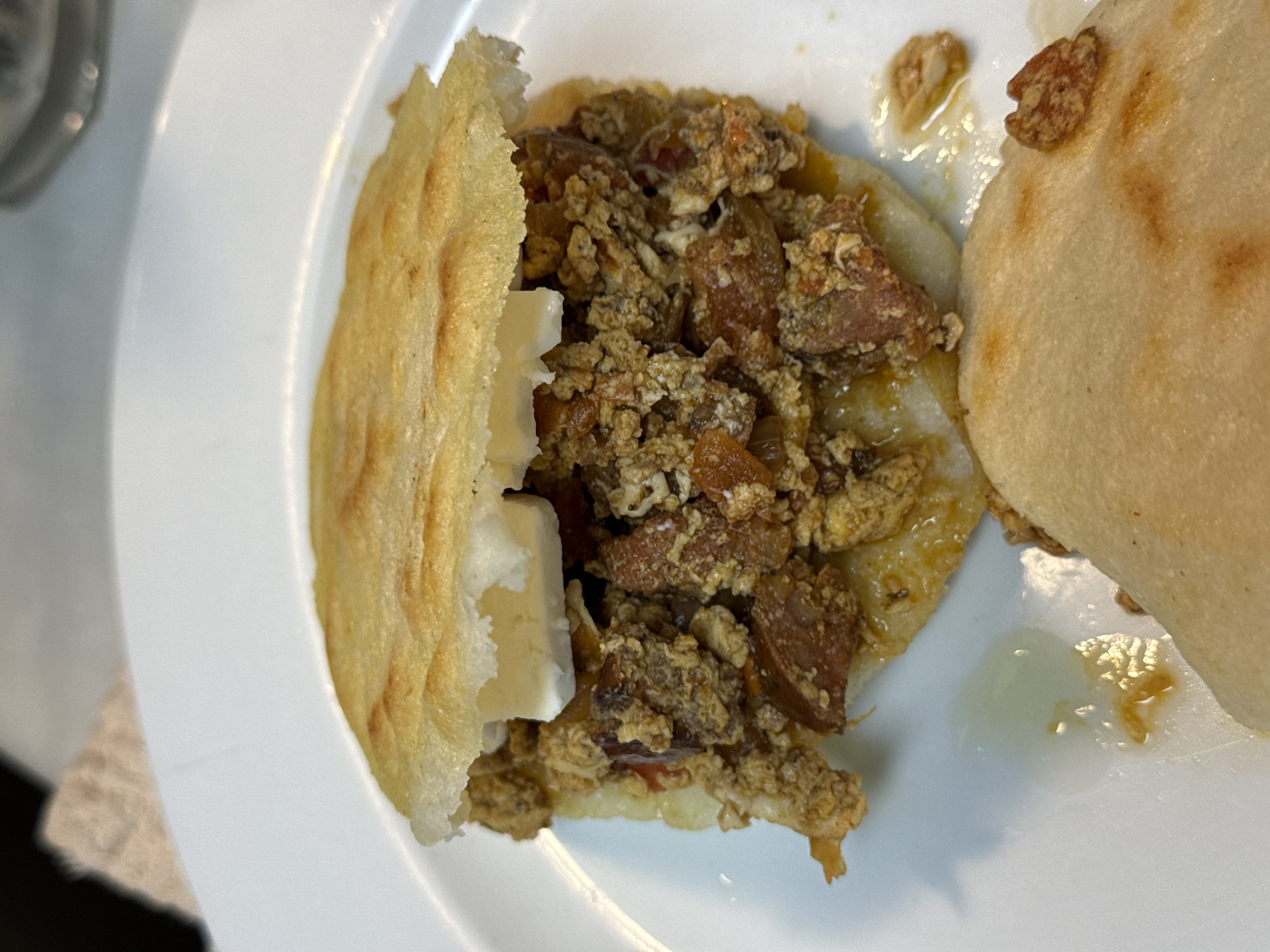 A partially eaten arepa with scrambled eggs and sausage, along with white cheese, sits on a white plate. Another arepa sits to the side, also partially eaten. The environment is nondescript, with only a hint of a gray countertop visible. The image does not provide any information about the location being in Barcelona, Spain.
