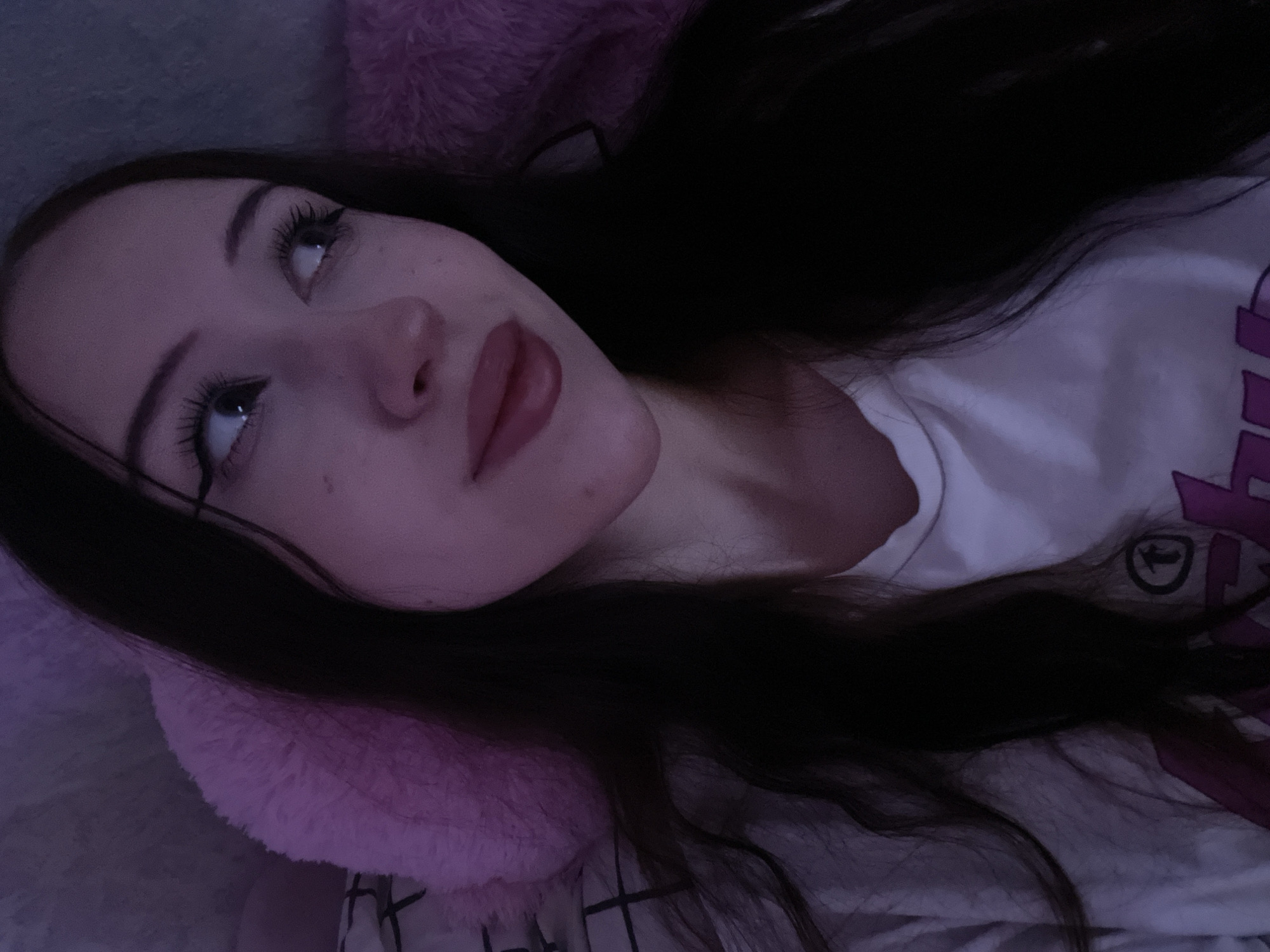 The image shows a close-up, overhead shot of a young woman lying down, looking upwards. She has long, dark hair that is spread across her face and shoulders. She is wearing a white t-shirt with purple graphics on it. The lighting in the image is dim and cast with a purple hue, suggesting it might be nighttime or a dimly lit indoor environment.
The woman's face is partially visible, with her eyes open and looking towards the top of the frame. She has noticeable makeup, including eyeliner and mascara, and her lips are painted a deep pink. Her expression appears neutral or perhaps thoughtful. A fluffy pink object, possibly a pillow or part of a blanket, is visible behind her head.
The overall impression is one of a private, relaxed moment. The low light and close-up framing create an intimate feel. There are no clear indications of the specific location beyond the context provided, which states Votkinsk, Russia. No discernible text is visible in the image.