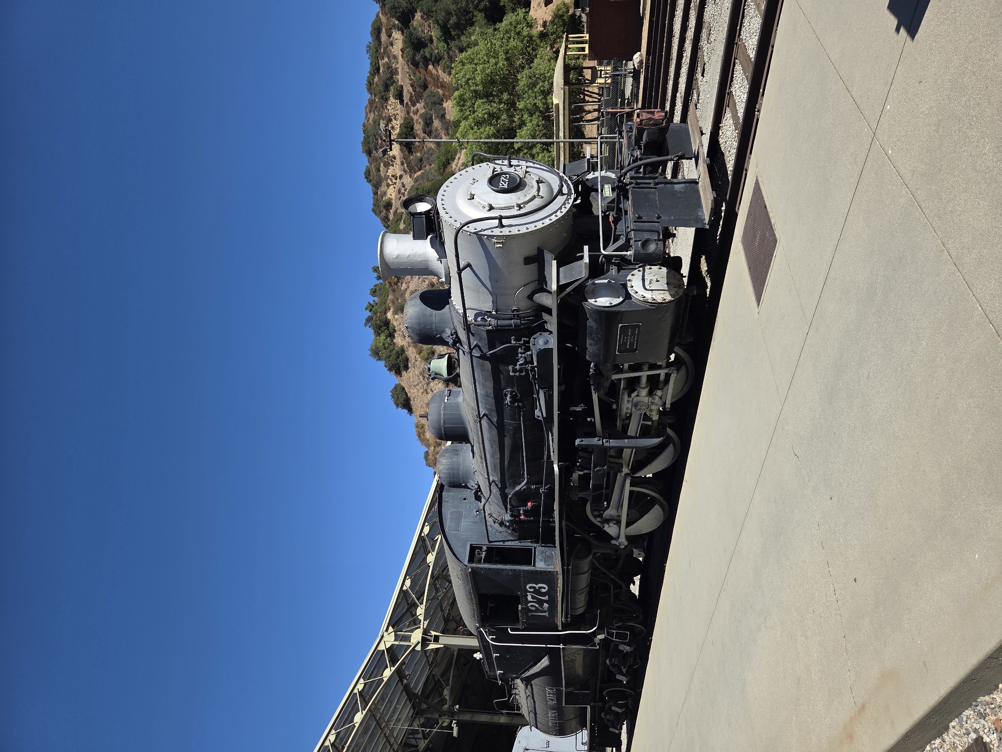 A vintage black steam locomotive, numbered "1273", is positioned on railroad tracks under a covered structure with a transparent roof. The locomotive has "SOUTHERN PACIFIC" written on its tender. To the right of the train, a grassy, hilly landscape with trees and a few structures is visible under a clear blue sky. The scene appears to be a museum or historical site, possibly the Travel Town Museum in Griffith Park, Los Angeles, California. There are no people present in the image.
