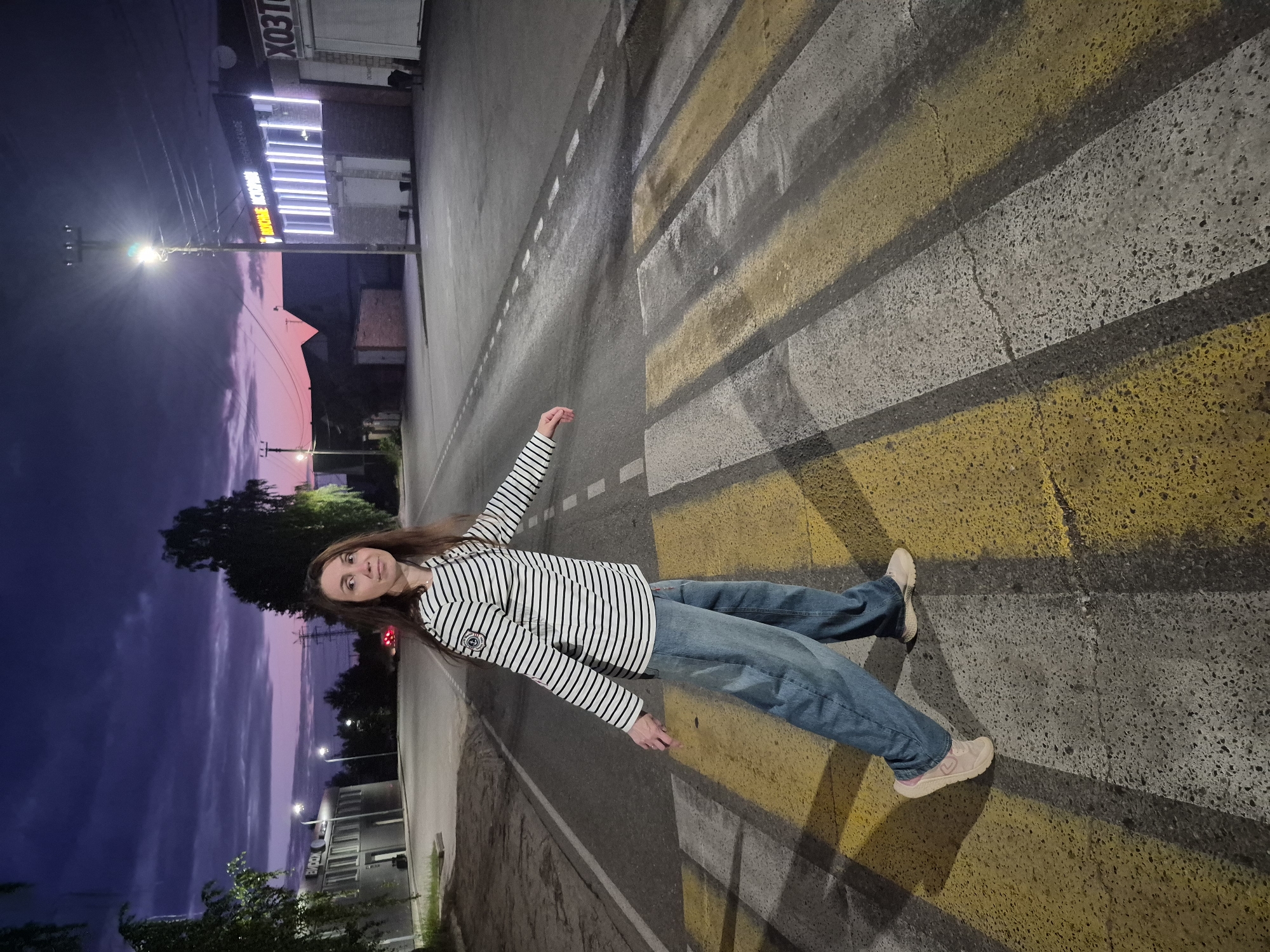 A woman with long brown hair is walking across a pedestrian crossing at dusk in the city of Chaikovsky, Russia. She is wearing a horizontally striped long-sleeved shirt and loose blue jeans. The sky is a deep purple with pink and orange hues near the horizon. In the background, there are commercial buildings with illuminated signs, streetlights, and trees. The pedestrian crossing has yellow and white stripes. The lighting suggests it is twilight, with artificial lights from the buildings and streetlights casting shadows.